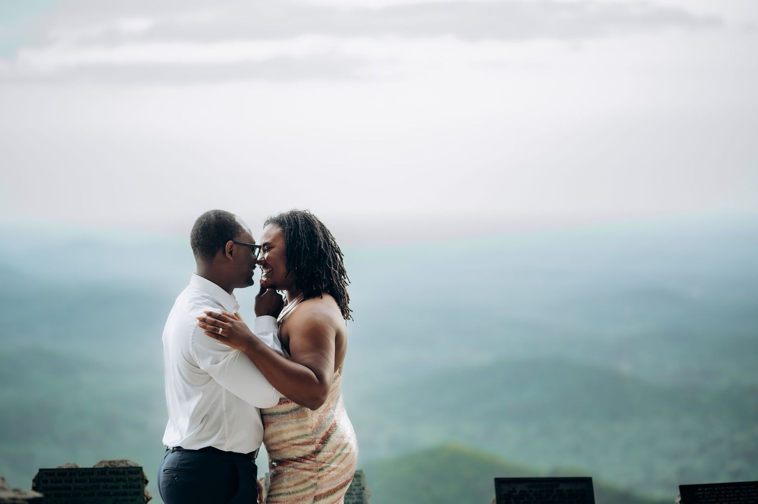 A couple embraces and touches foreheads on a scenic overlook with misty mountain views in the background.