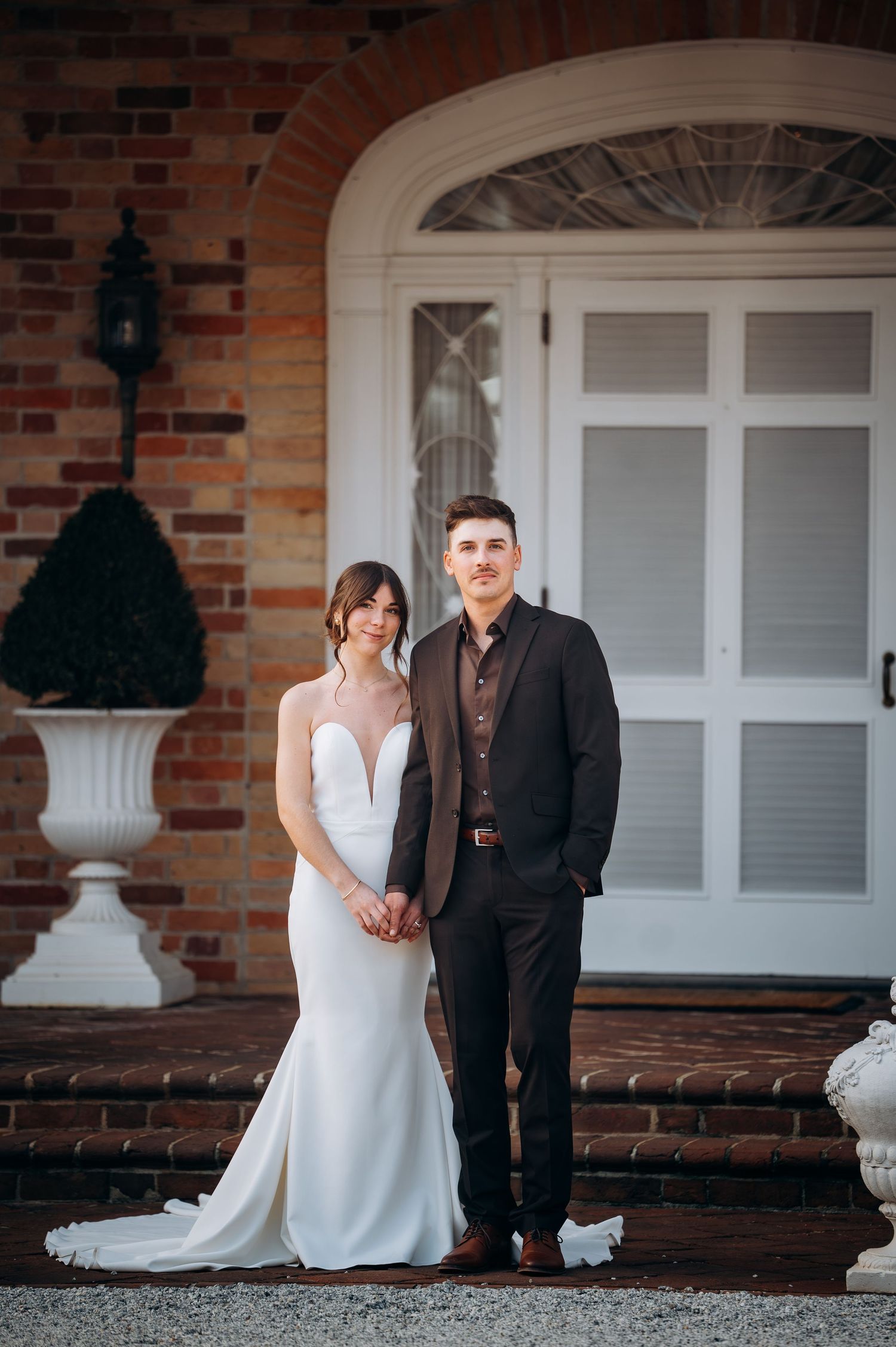 Elegant couple poses in front of a classic brick entrance with white trim and windows.