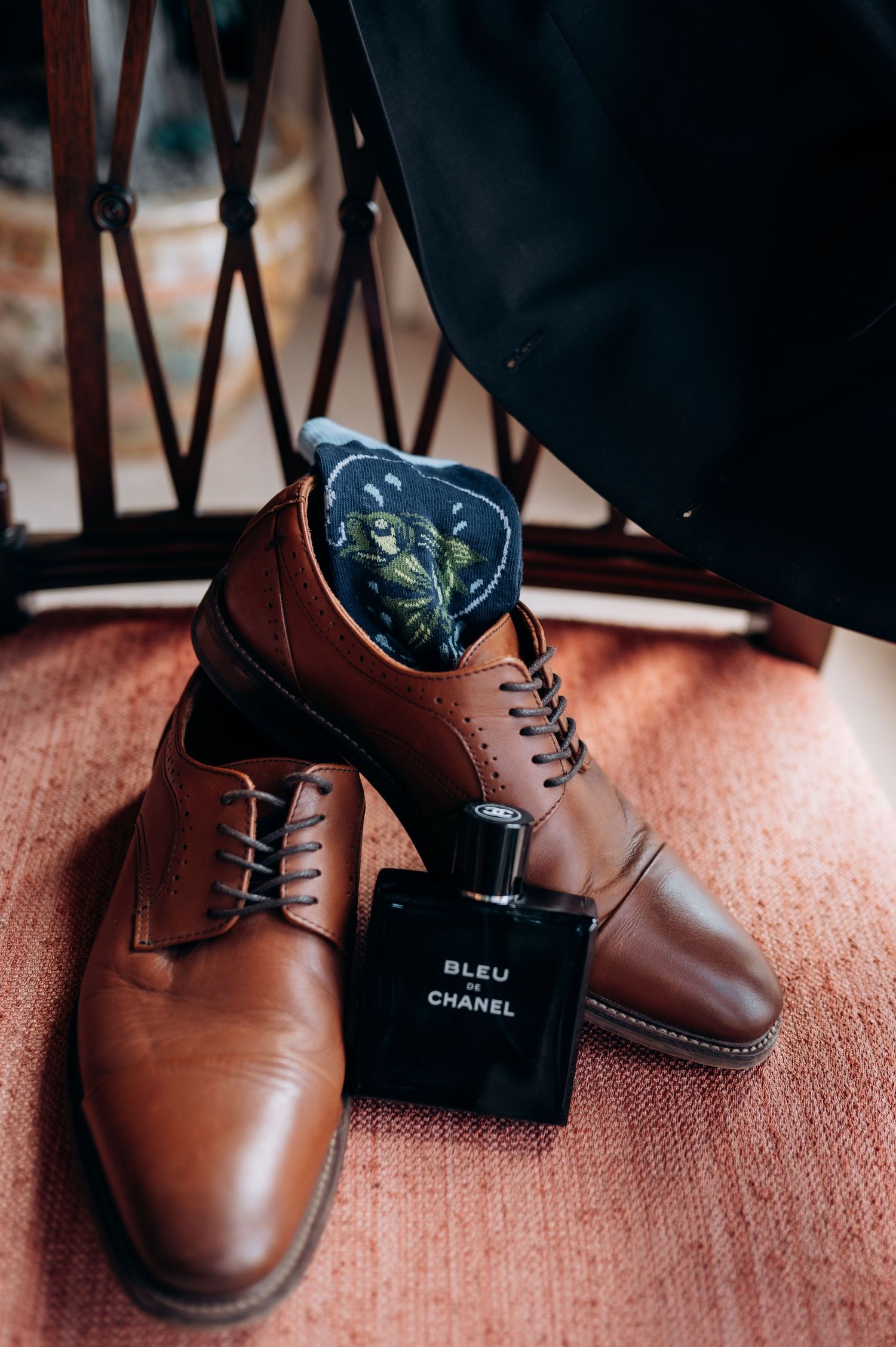 Close-up of brown leather dress shoes with Chanel Bleu fragrance bottle on reddish carpet.