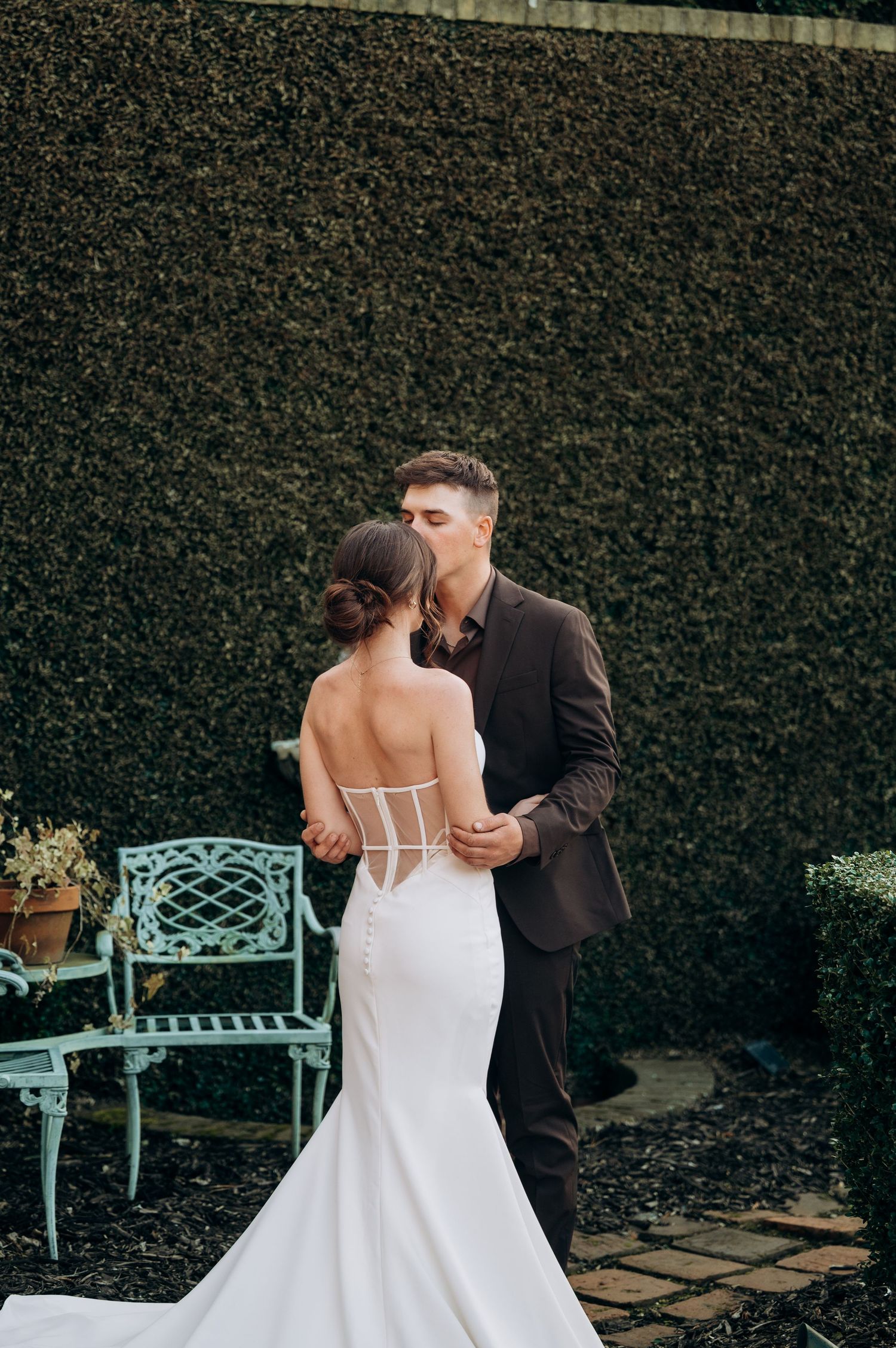 Intimate wedding moment captured against lush garden hedge with vintage turquoise bench.