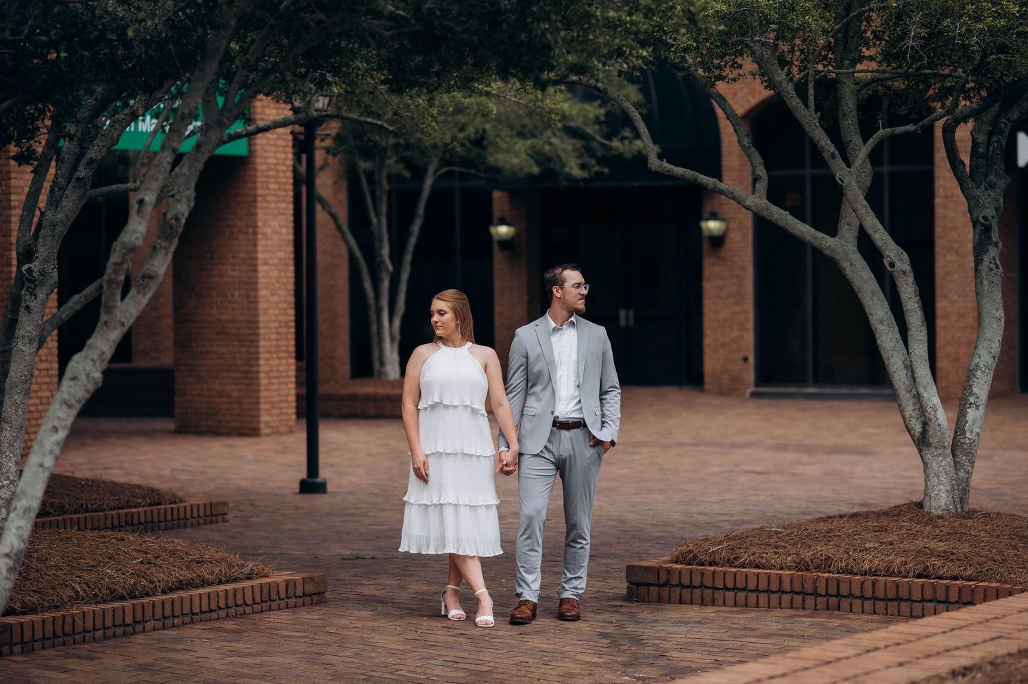 Couple walking together along tree-lined brick courtyard of historic building.