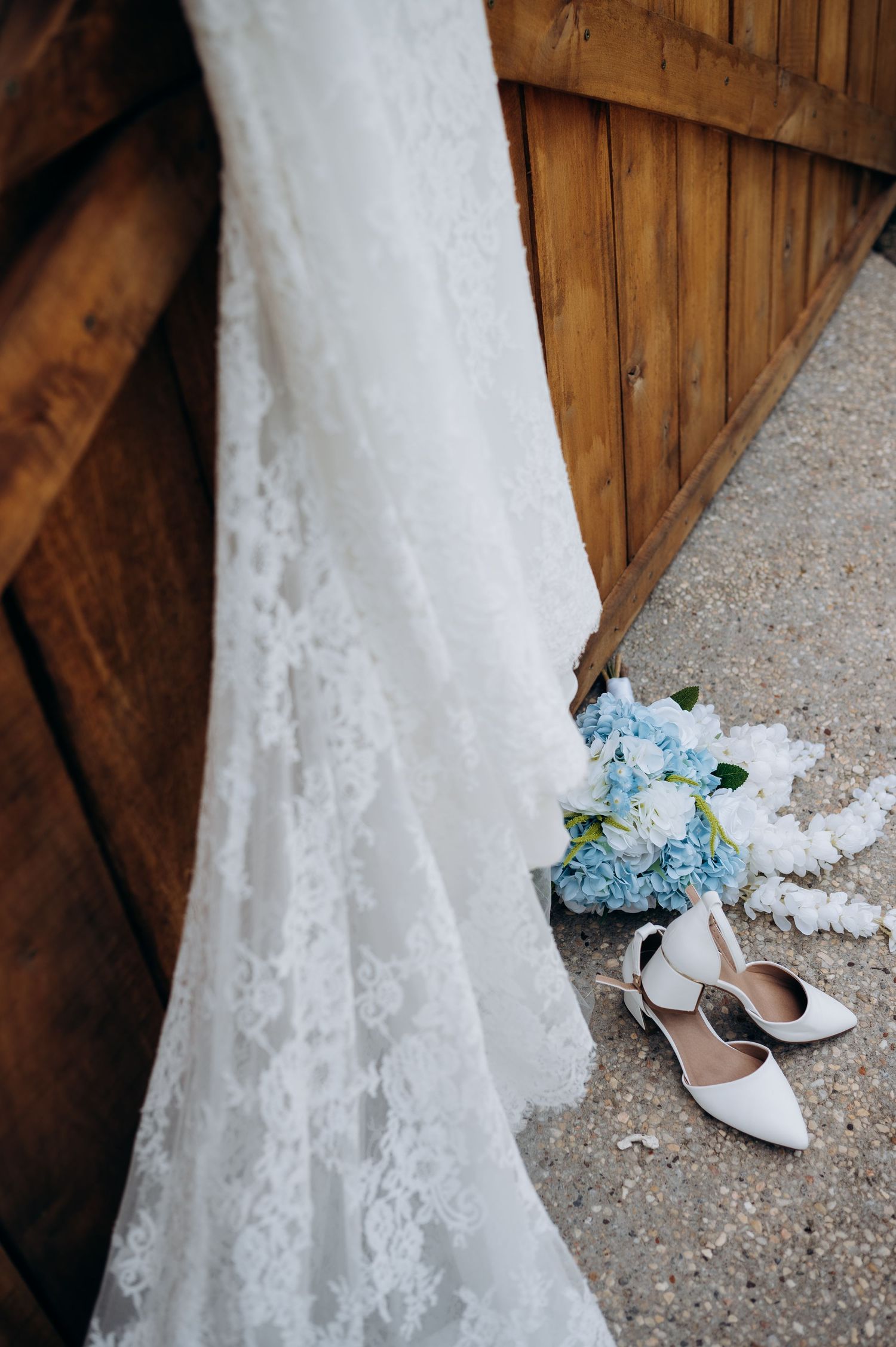 White lace wedding dress hanging near wooden barn door with blue hydrangea bouquet and white bridal heels on sandy ground.