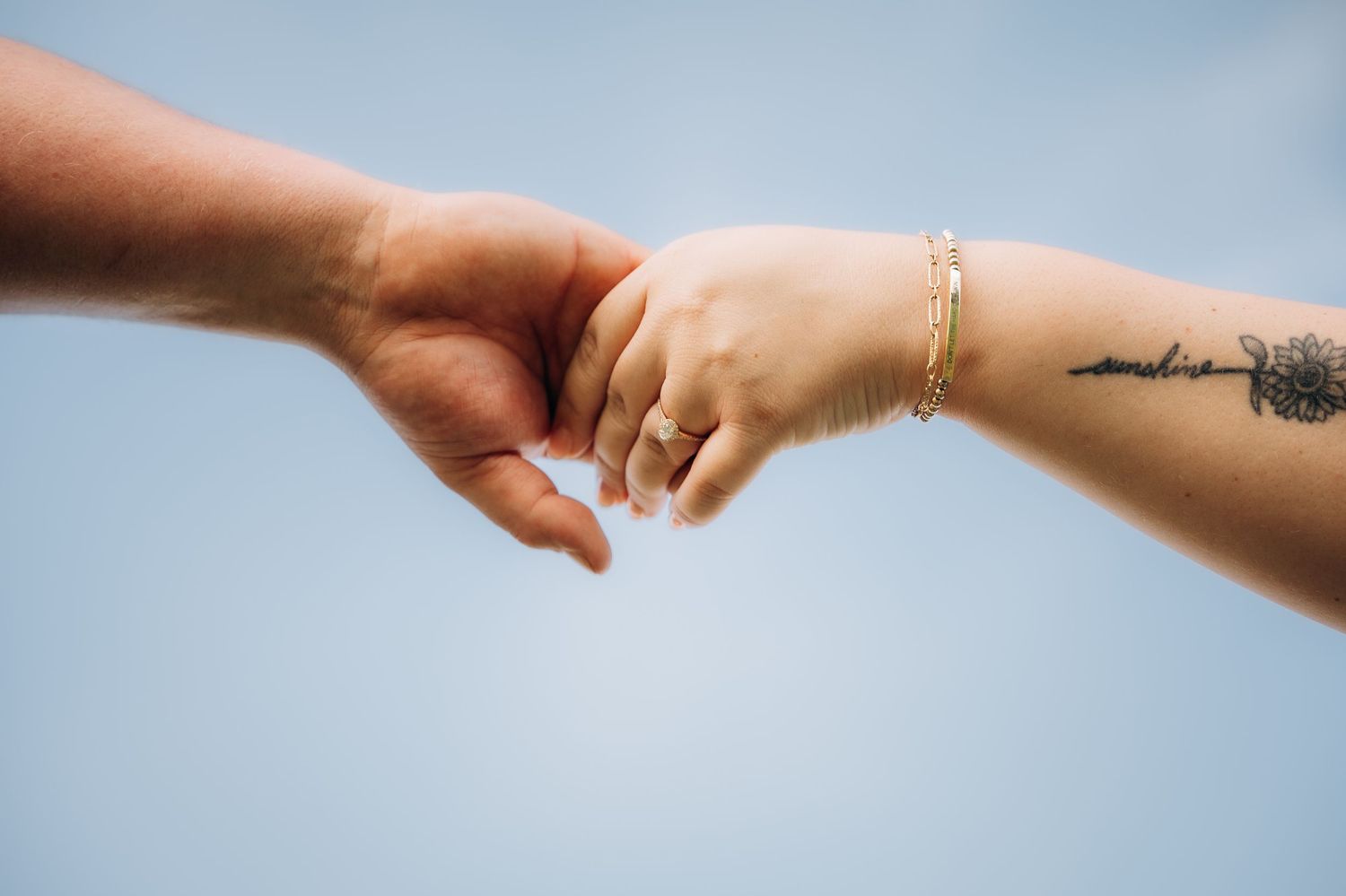 Two hands gently touching fingertips against a blue sky, one with a tattoo and gold bracelet.