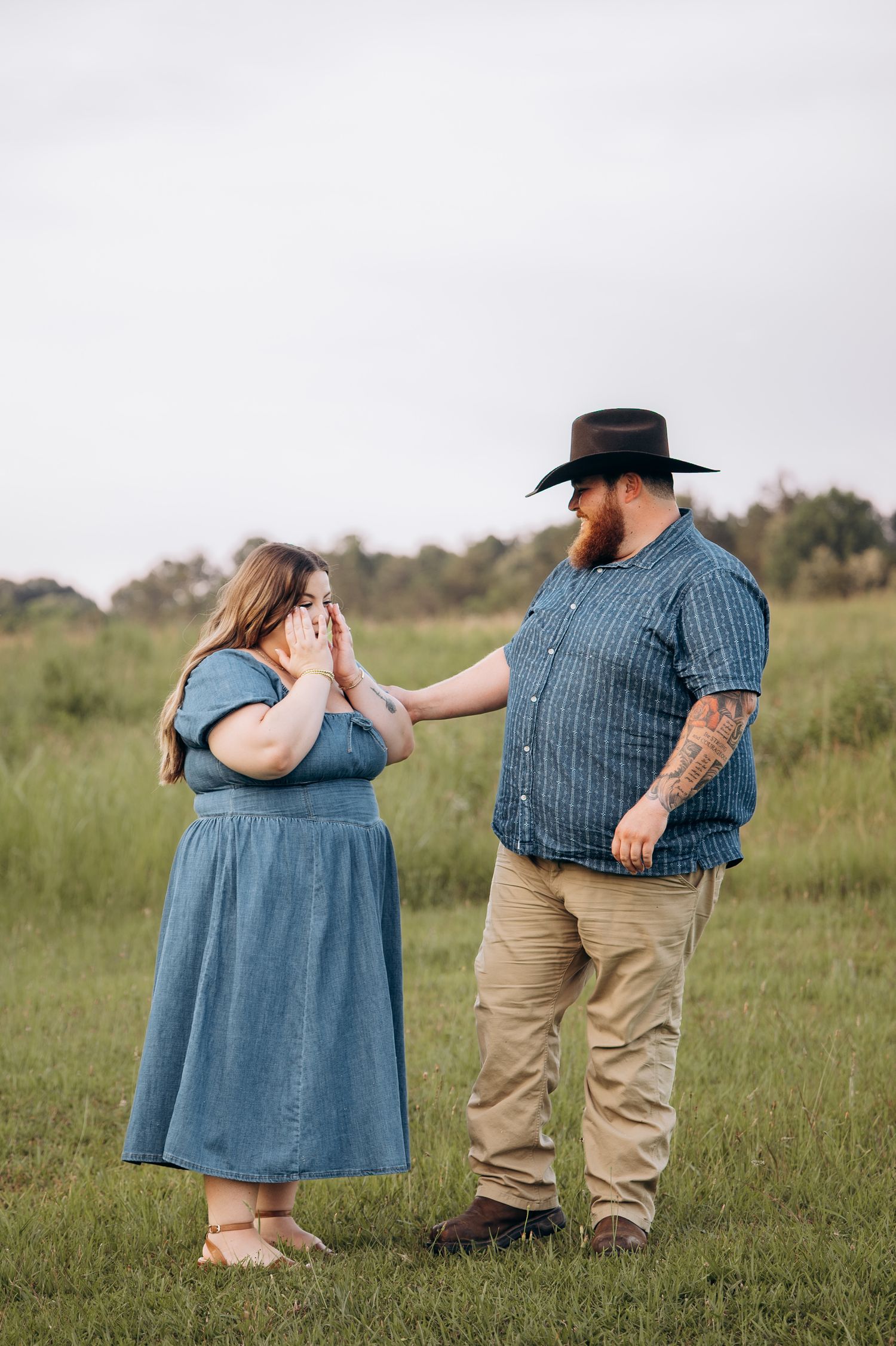Man in cowboy hat comforting tearful woman in blue dress standing in a green meadow outdoors.