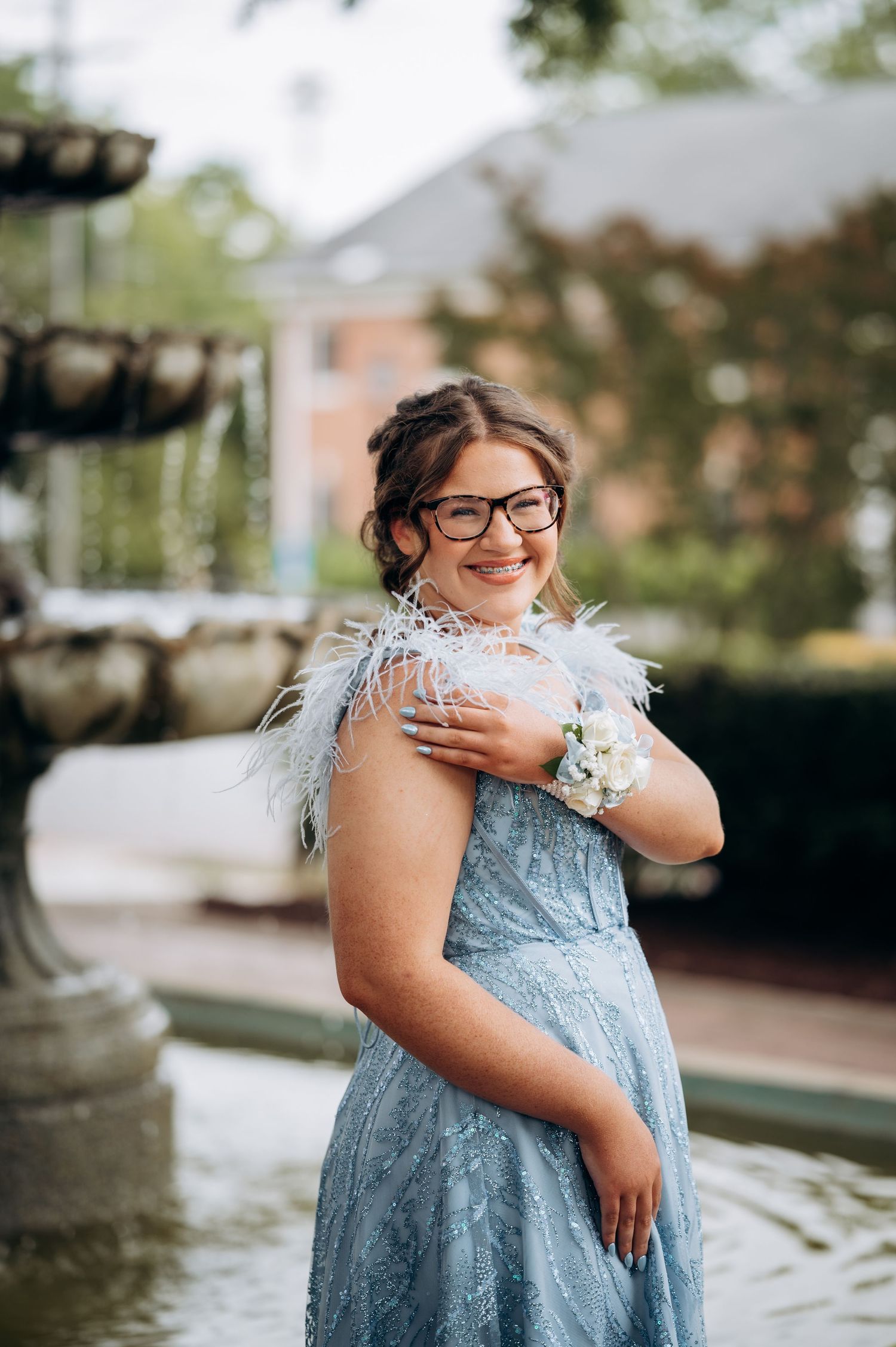 Young woman in blue feathered prom dress with wrist corsage smiling near a fountain.