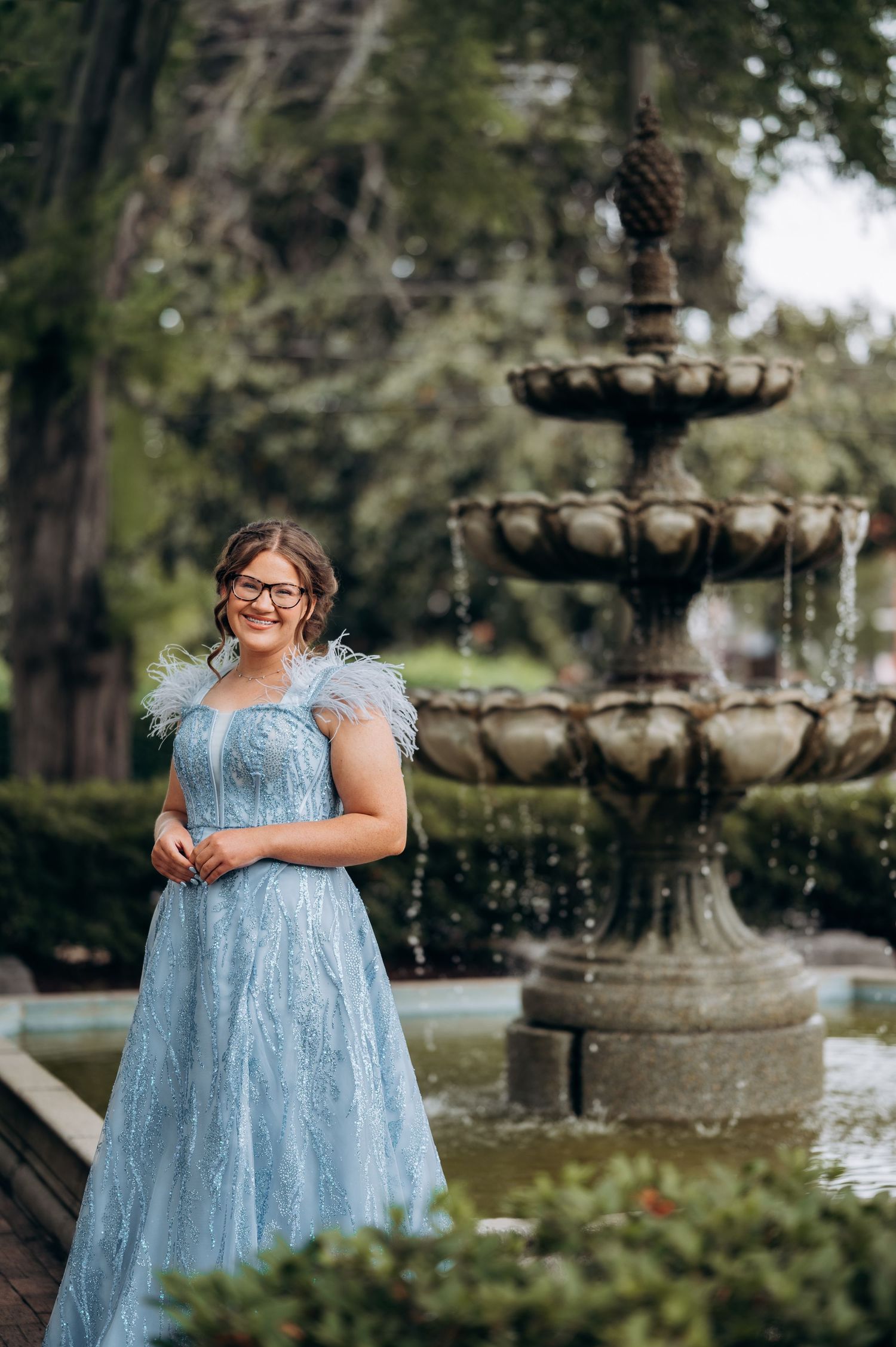 Young woman in blue feathered gown smiling by stone fountain in garden.