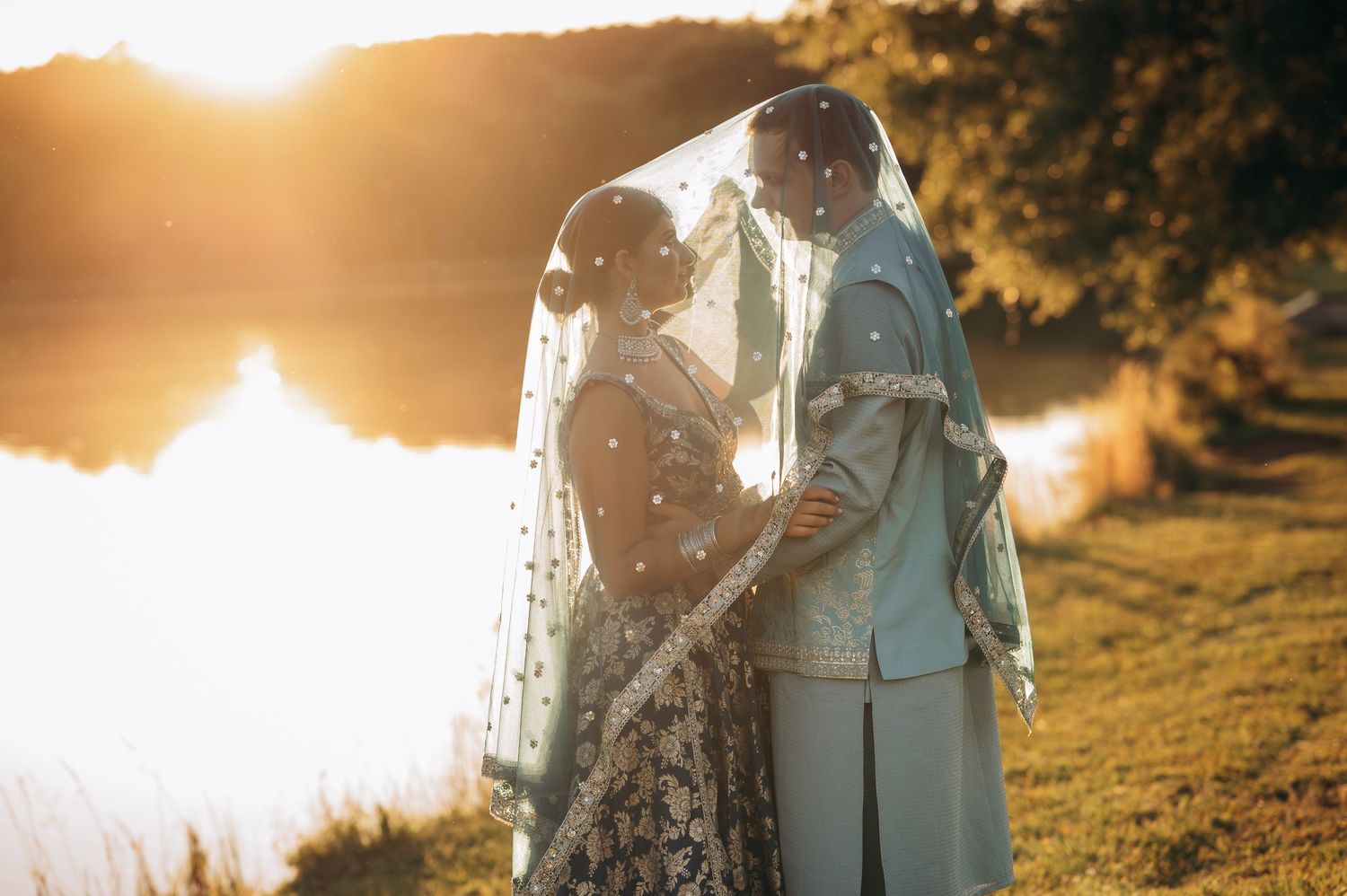 Indian bride and groom sharing romantic moment under embroidered veil with stunning golden sunset lake backdrop.