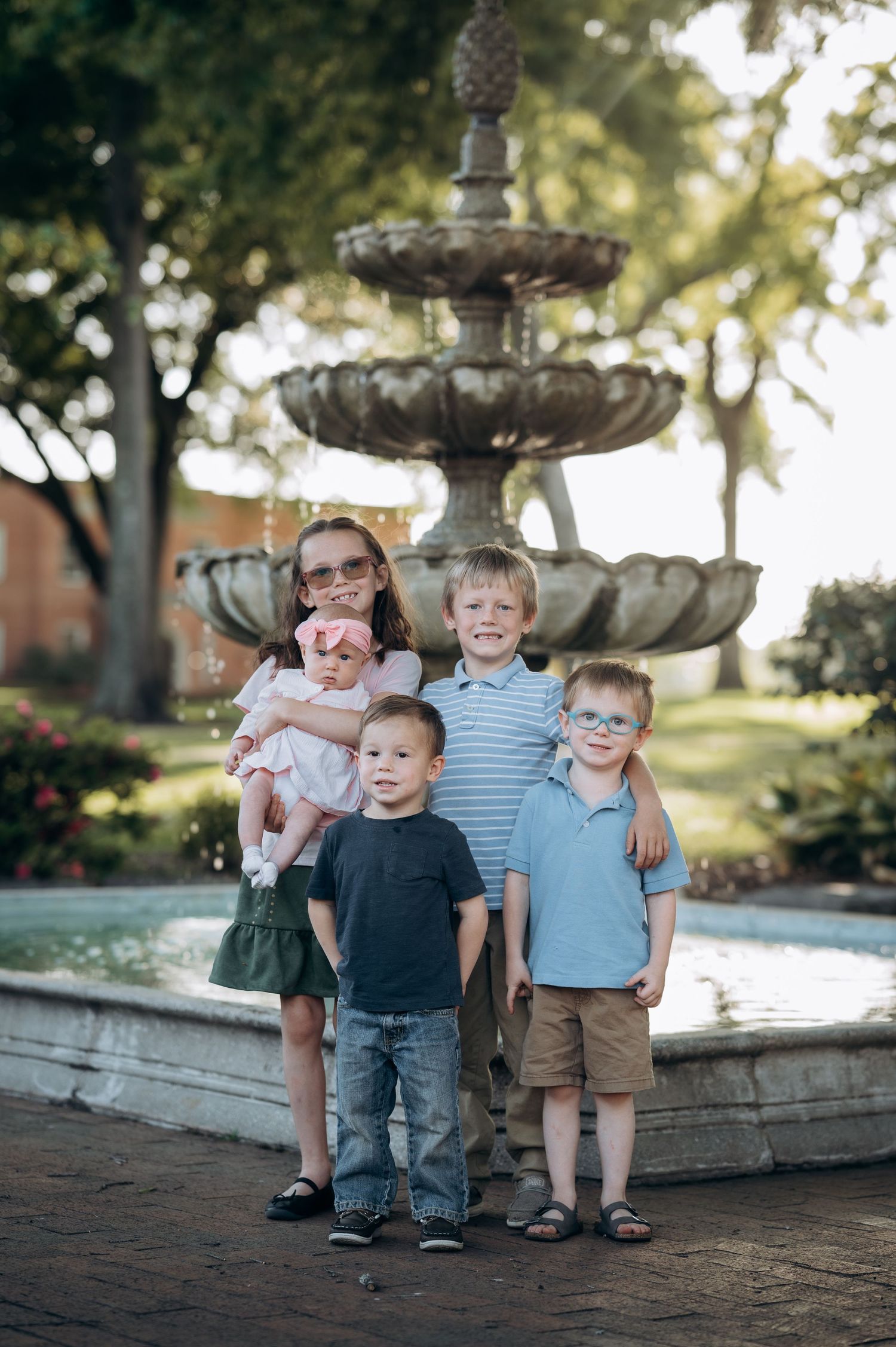 Four children pose in front of a tiered stone fountain in a park, with the oldest girl holding a baby.