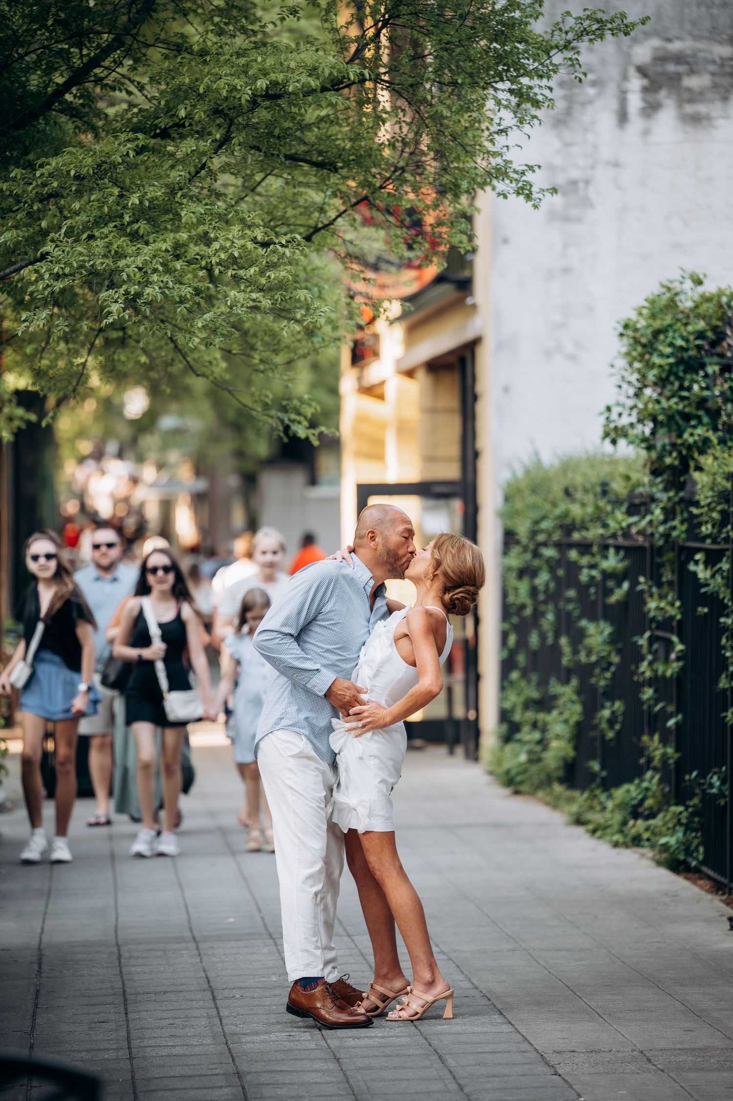 Couple sharing a passionate kiss on a busy sidewalk, man in blue shirt and woman in white dress.