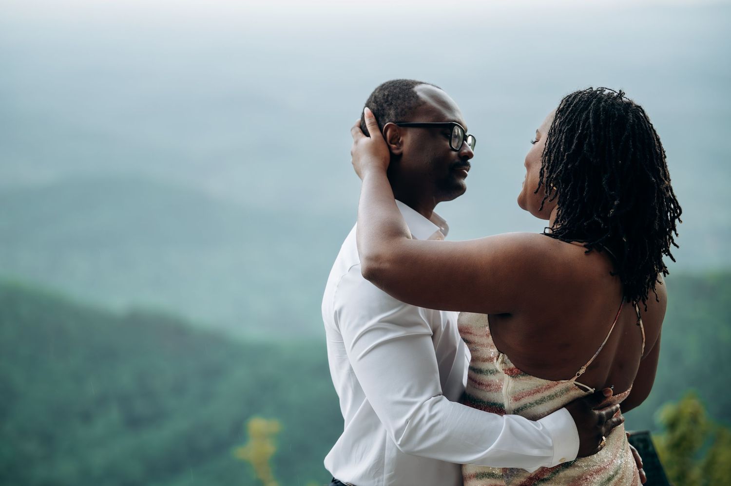 A Black couple shares a romantic embrace on a scenic mountain overlook, the woman tenderly holding the mans face.