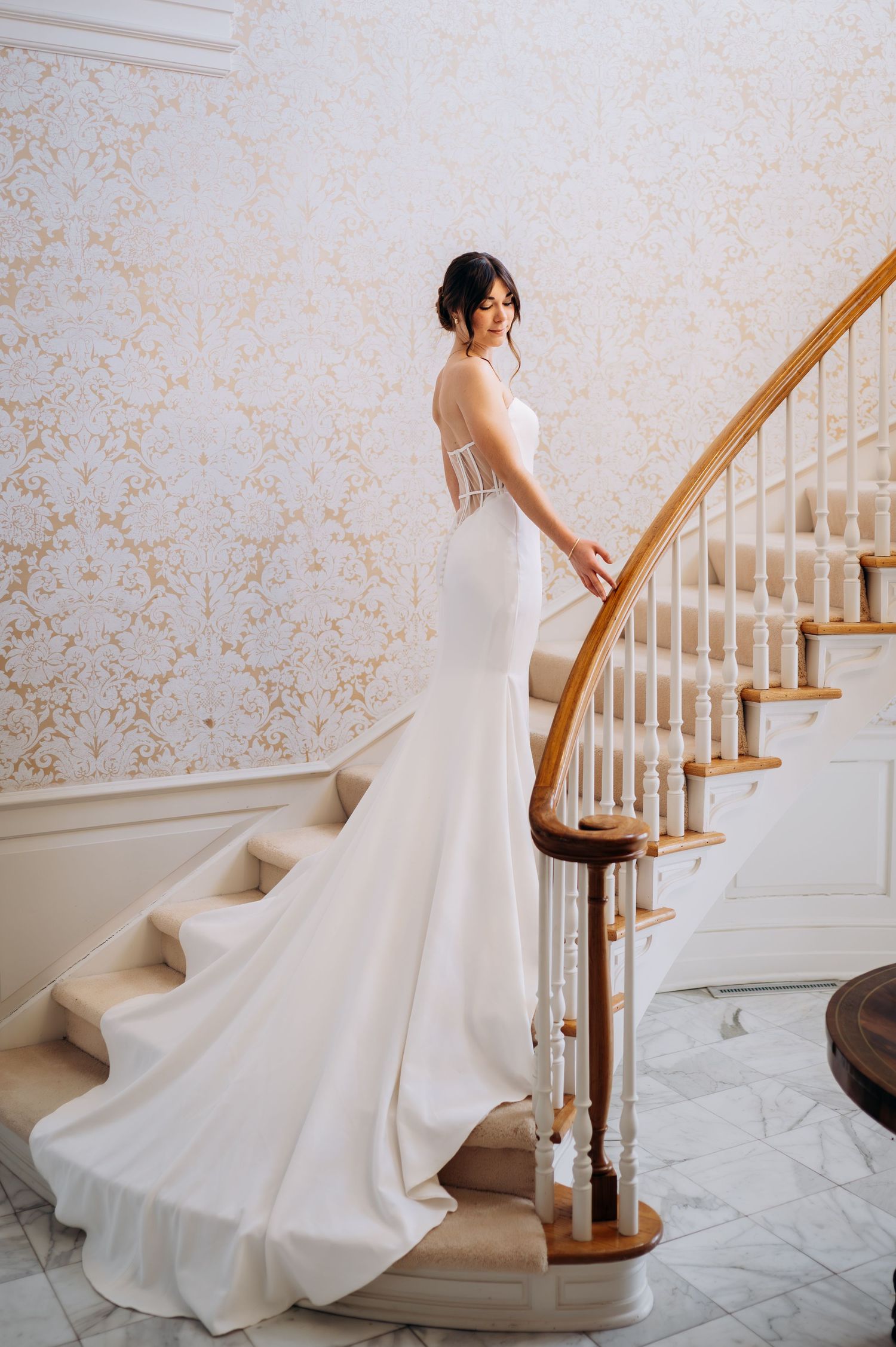 Bride in elegant mermaid wedding gown with sheer back poses on grand staircase with ornate wallpaper.