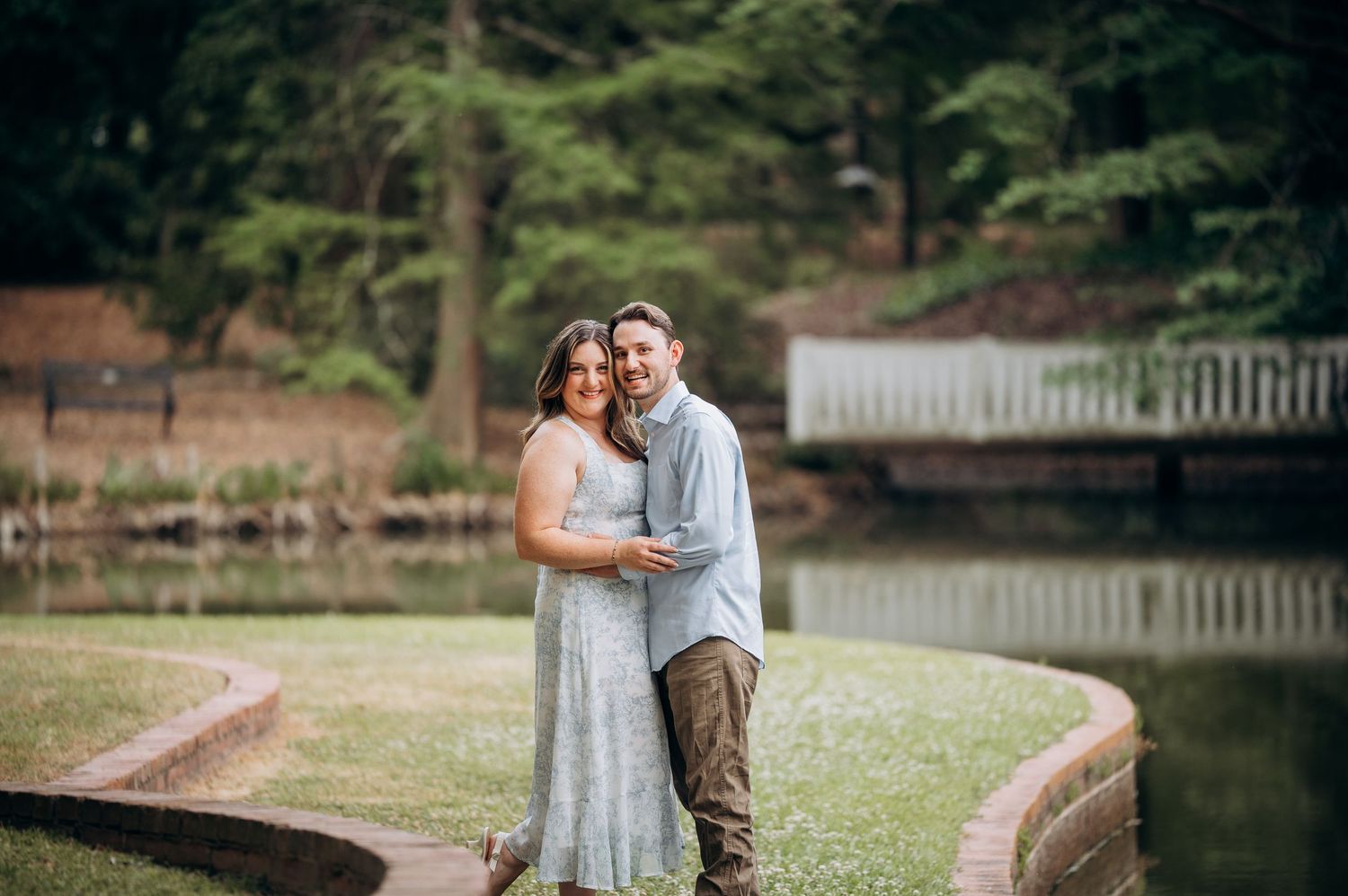 Couple smiling and embracing on a curved brick path beside a peaceful garden pond.