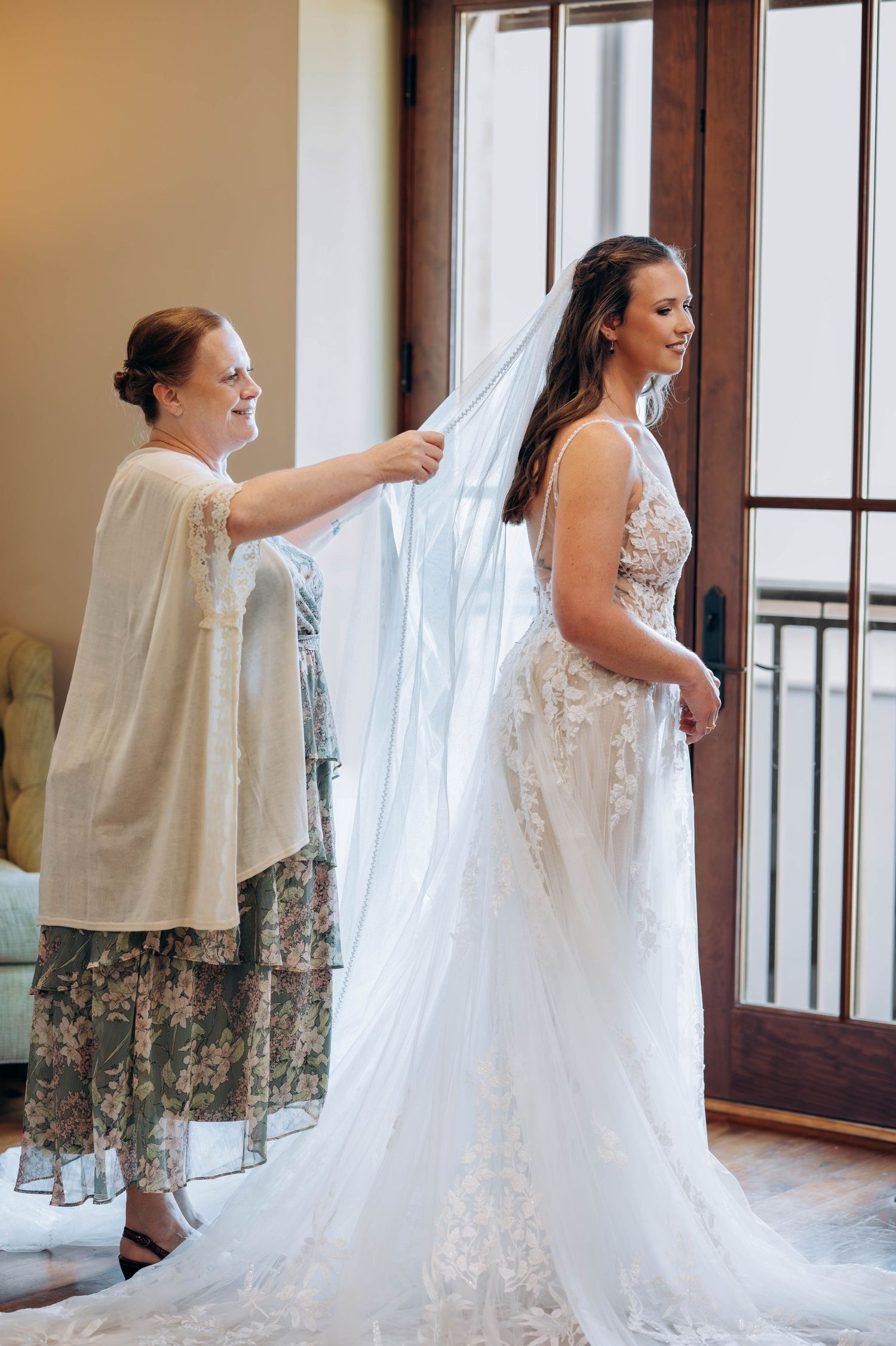 A flowing white lace wedding dress being fitted as sunlight streams through French doors.