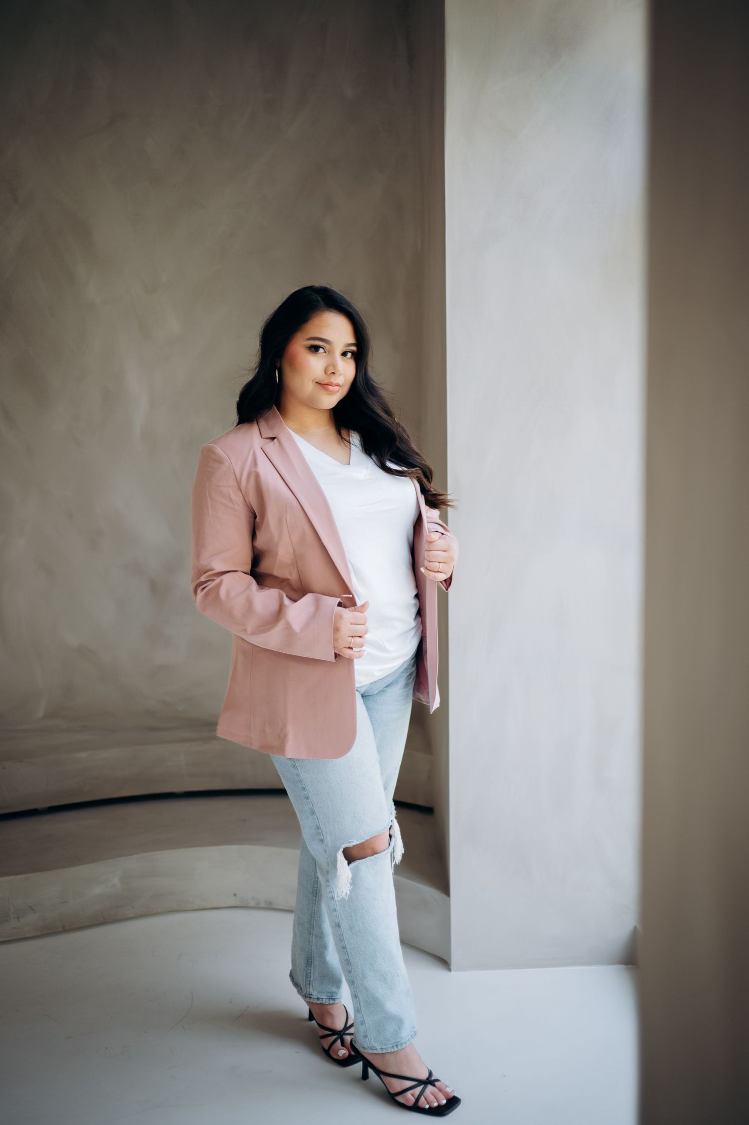 Fashion model wearing pink blazer, white top and distressed jeans poses against textured grey wall.