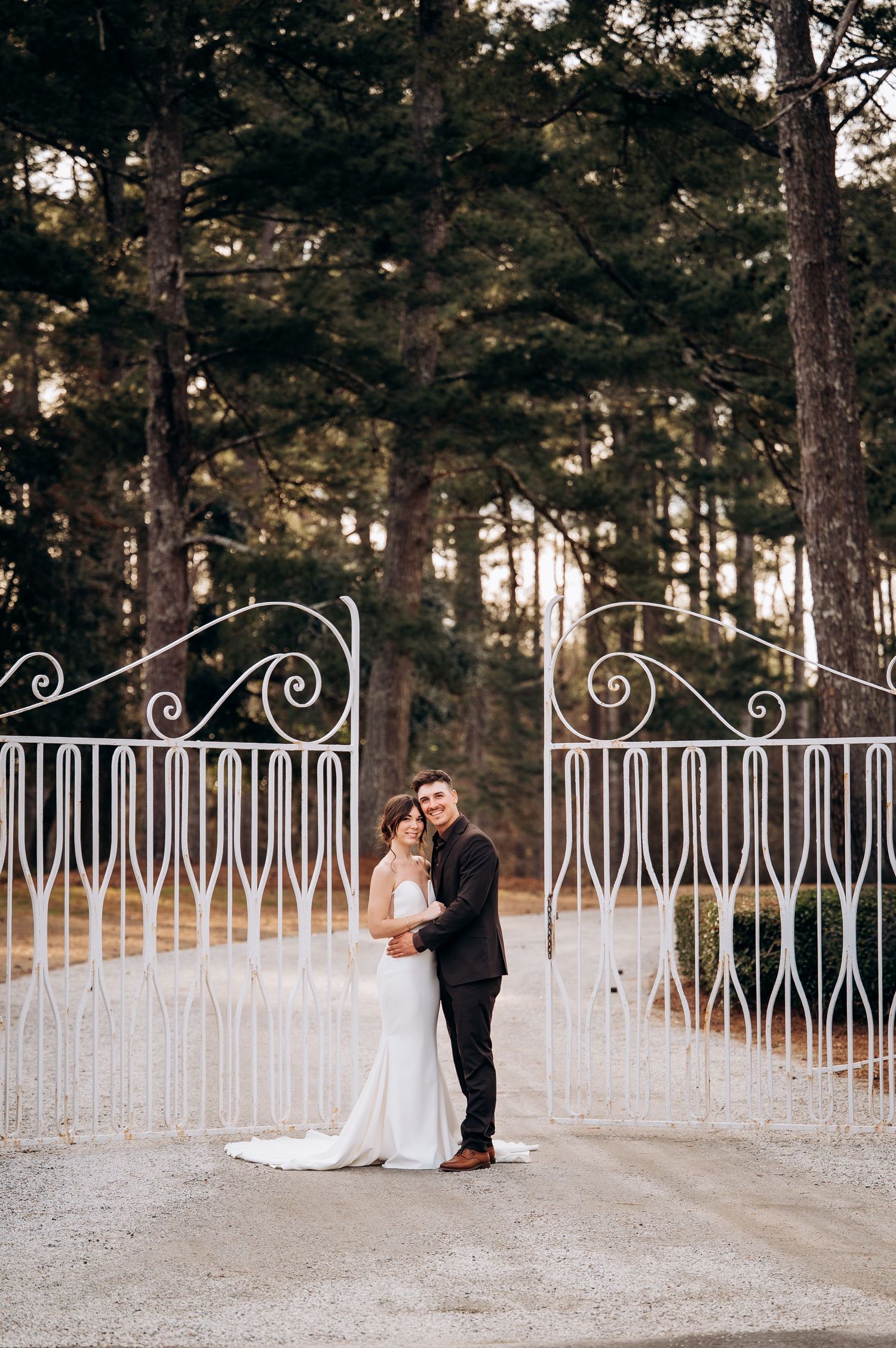 Bride and groom embrace before ornate white iron gates surrounded by tall pine trees at golden hour.