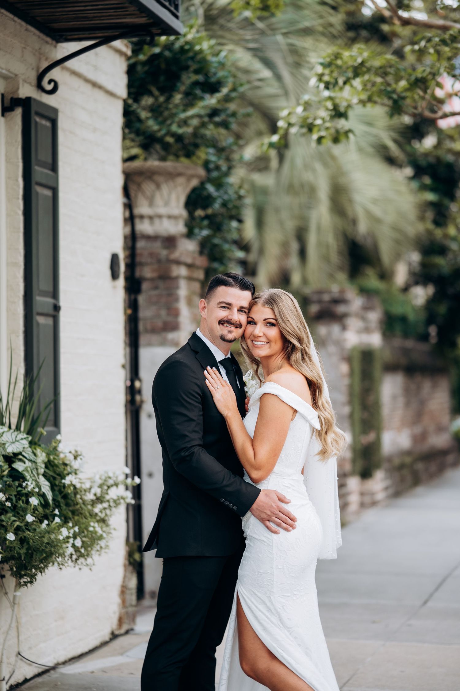 Bride in white gown and groom in black tuxedo embracing on a charming city street.
