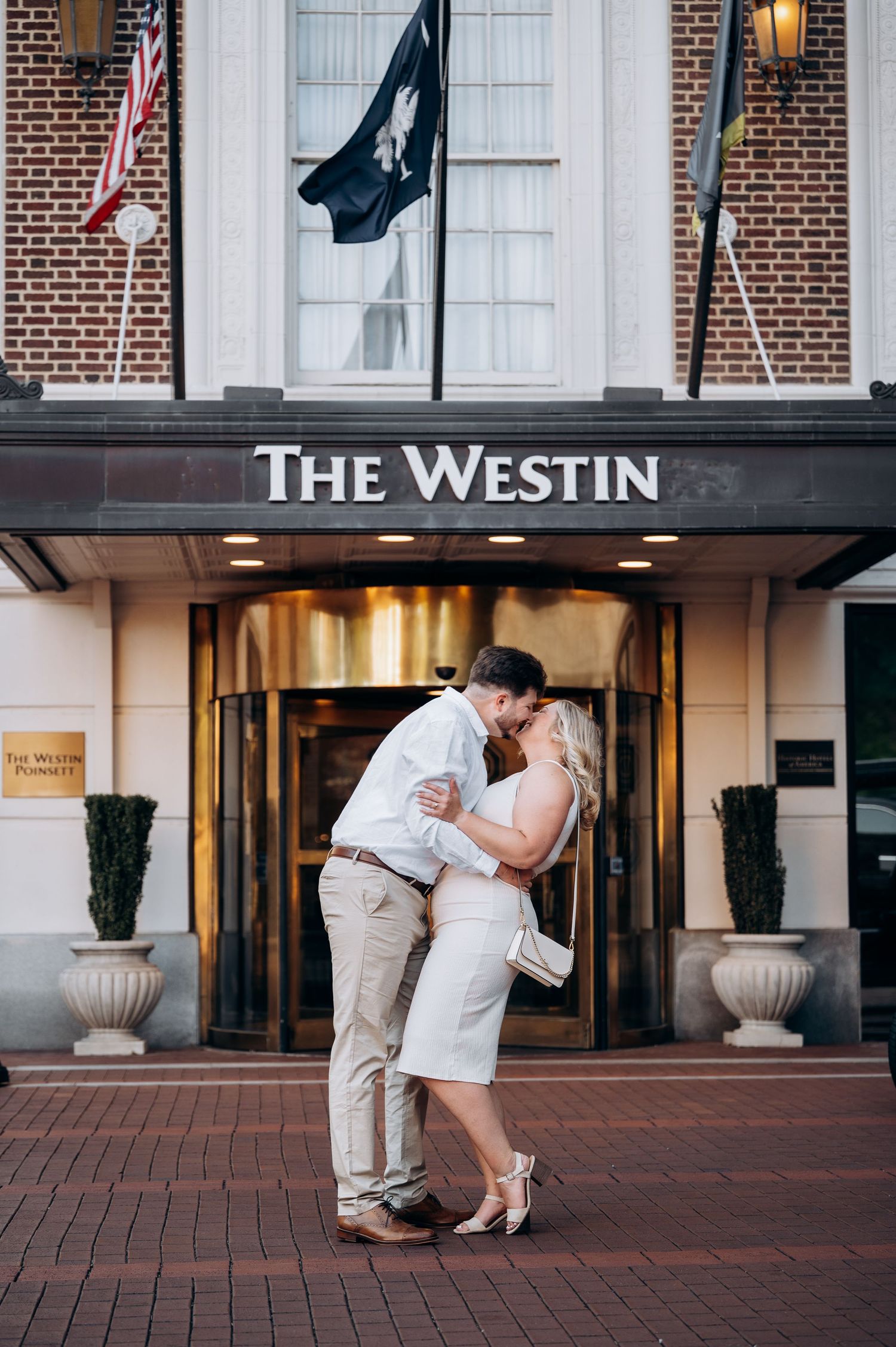 Couple kissing in front of The Westin hotel entrance on a brick plaza with flags overhead.