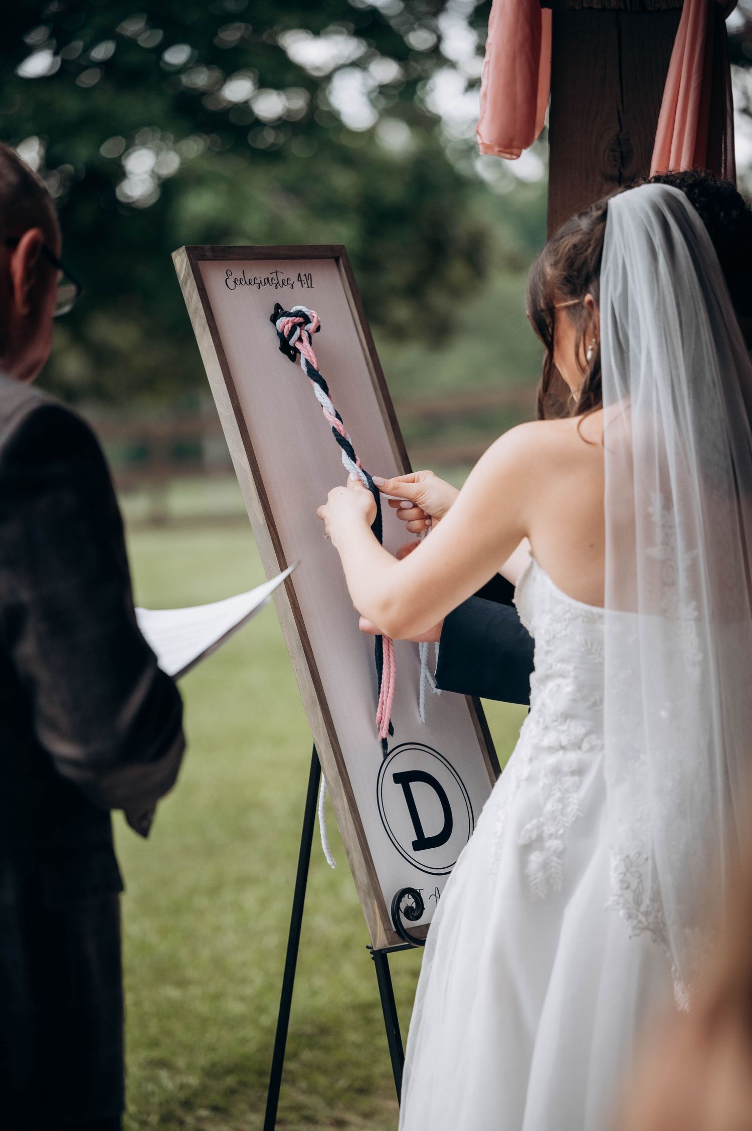 Bride in white dress signing a decorative board during an outdoor wedding ceremony.