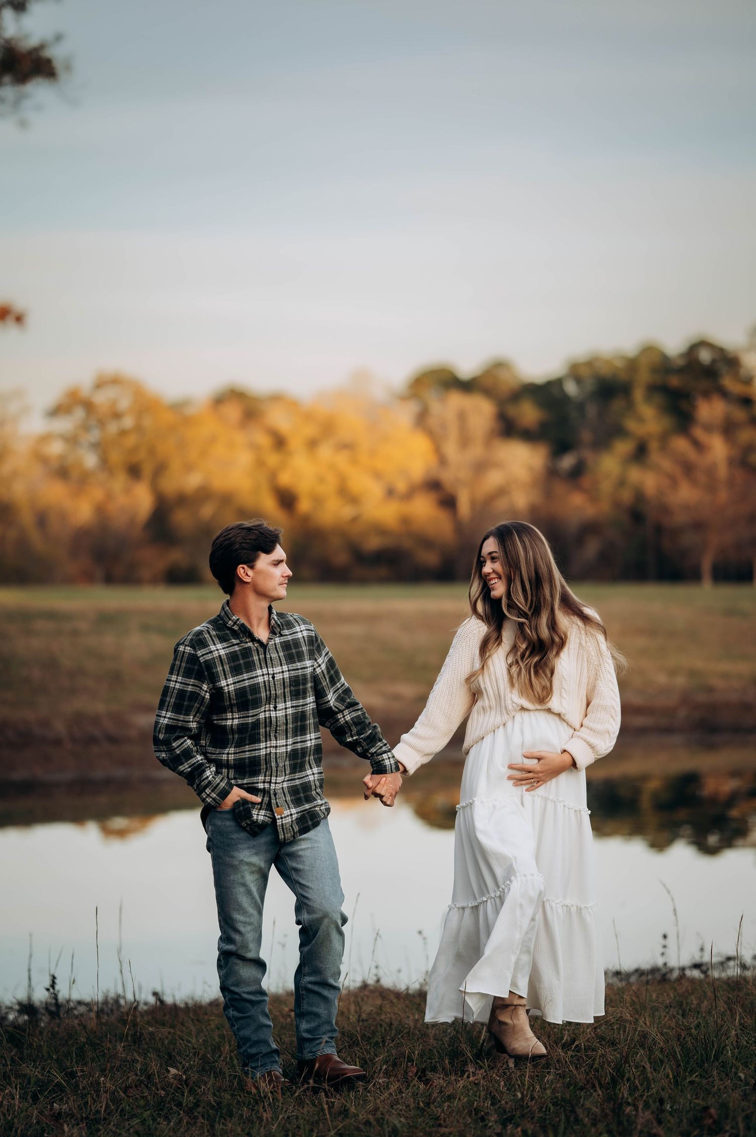 A couple stands by a pond at sunset holding hands while wearing casual fall attire.