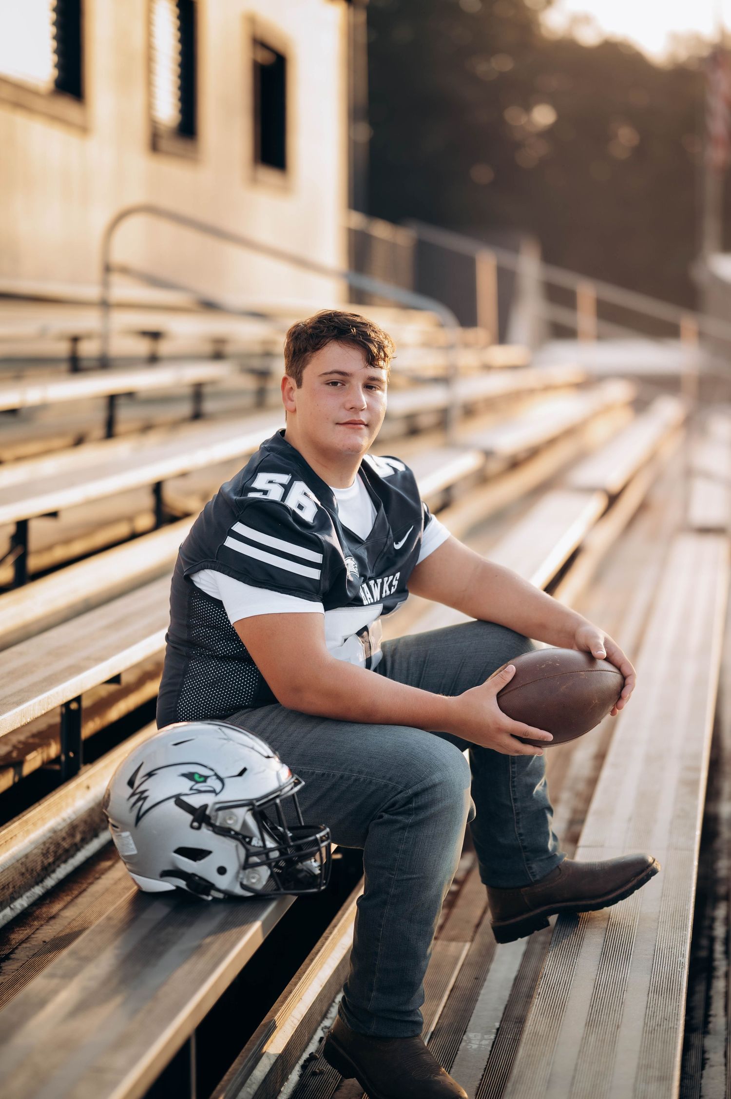 Football player in jersey sits thoughtfully on stadium bleachers during sunset.