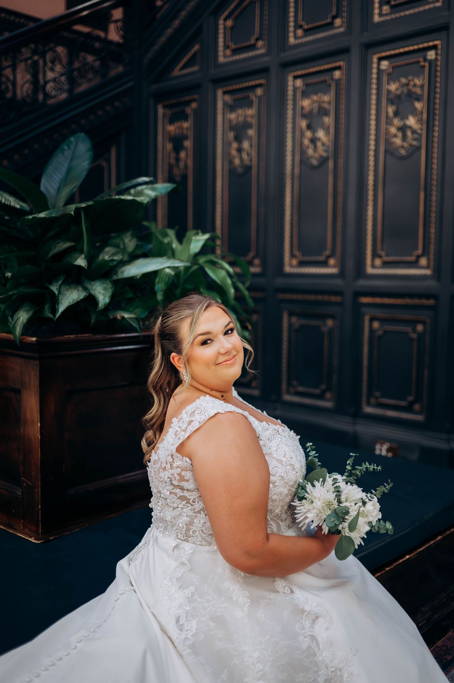 Bride in lace detailed wedding gown poses against dark paneled wall.