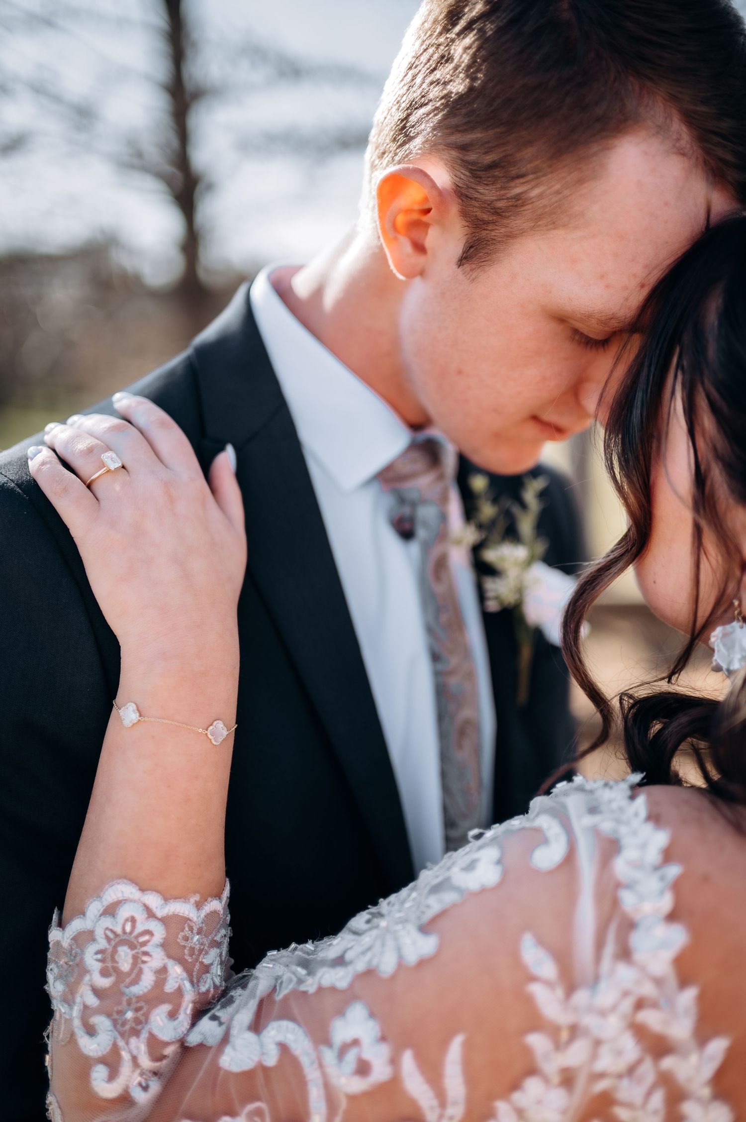 Bride in lace gown rests hand on groom's shoulder in tender wedding portrait outdoors.