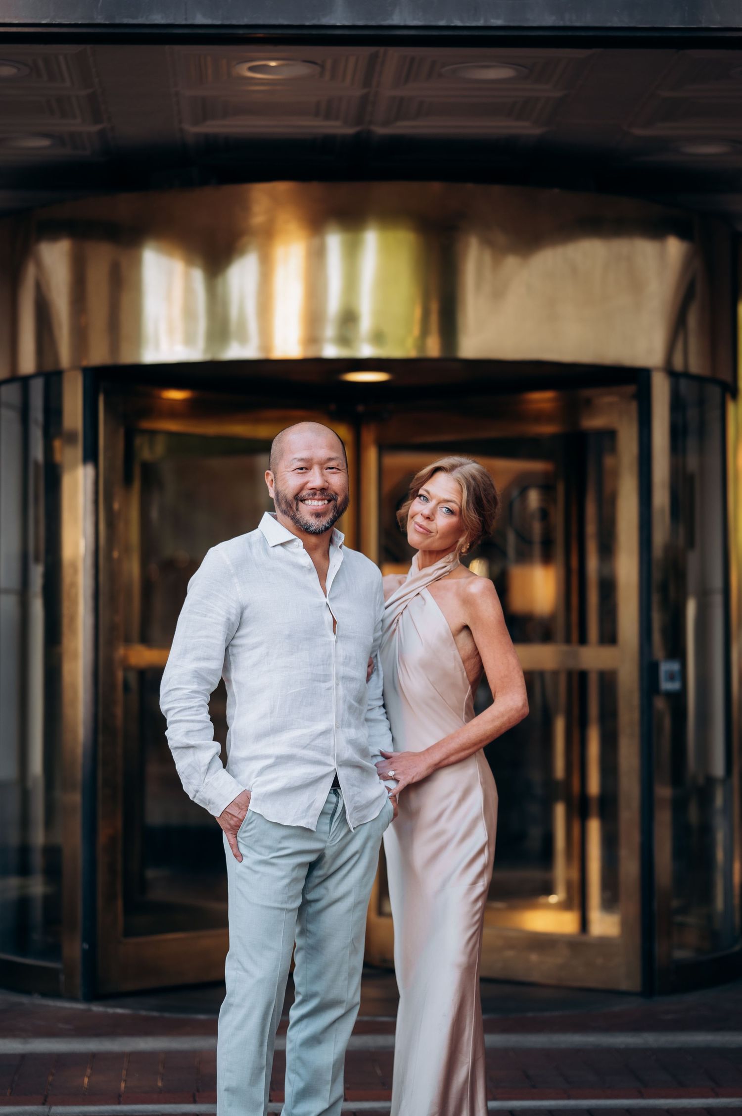 Stylish couple posing outside luxury hotel revolving door, man in white linen suit, woman in champagne satin gown.