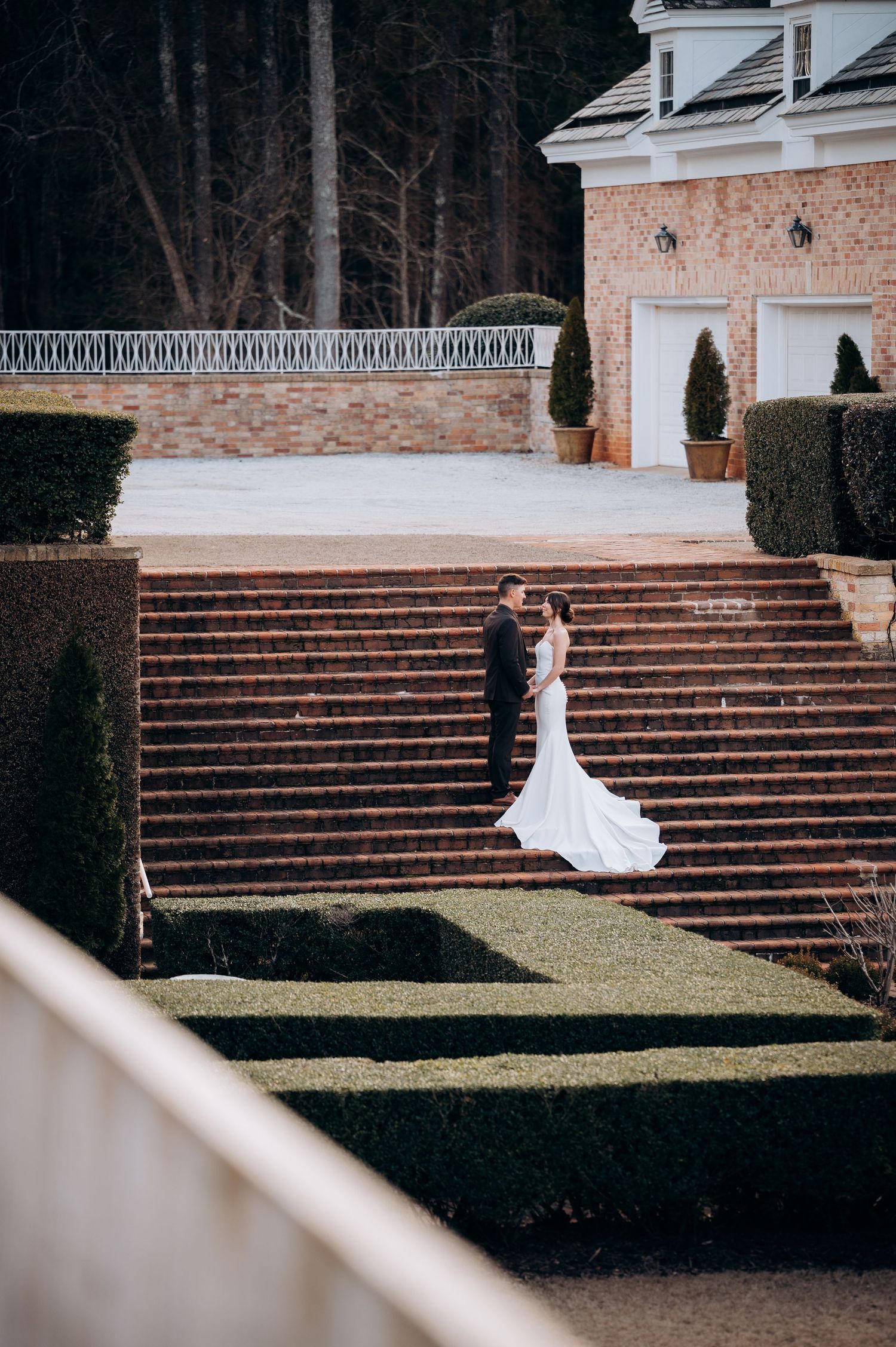 Bride and groom share intimate moment on grand brick staircase surrounded by manicured hedges at estate venue.