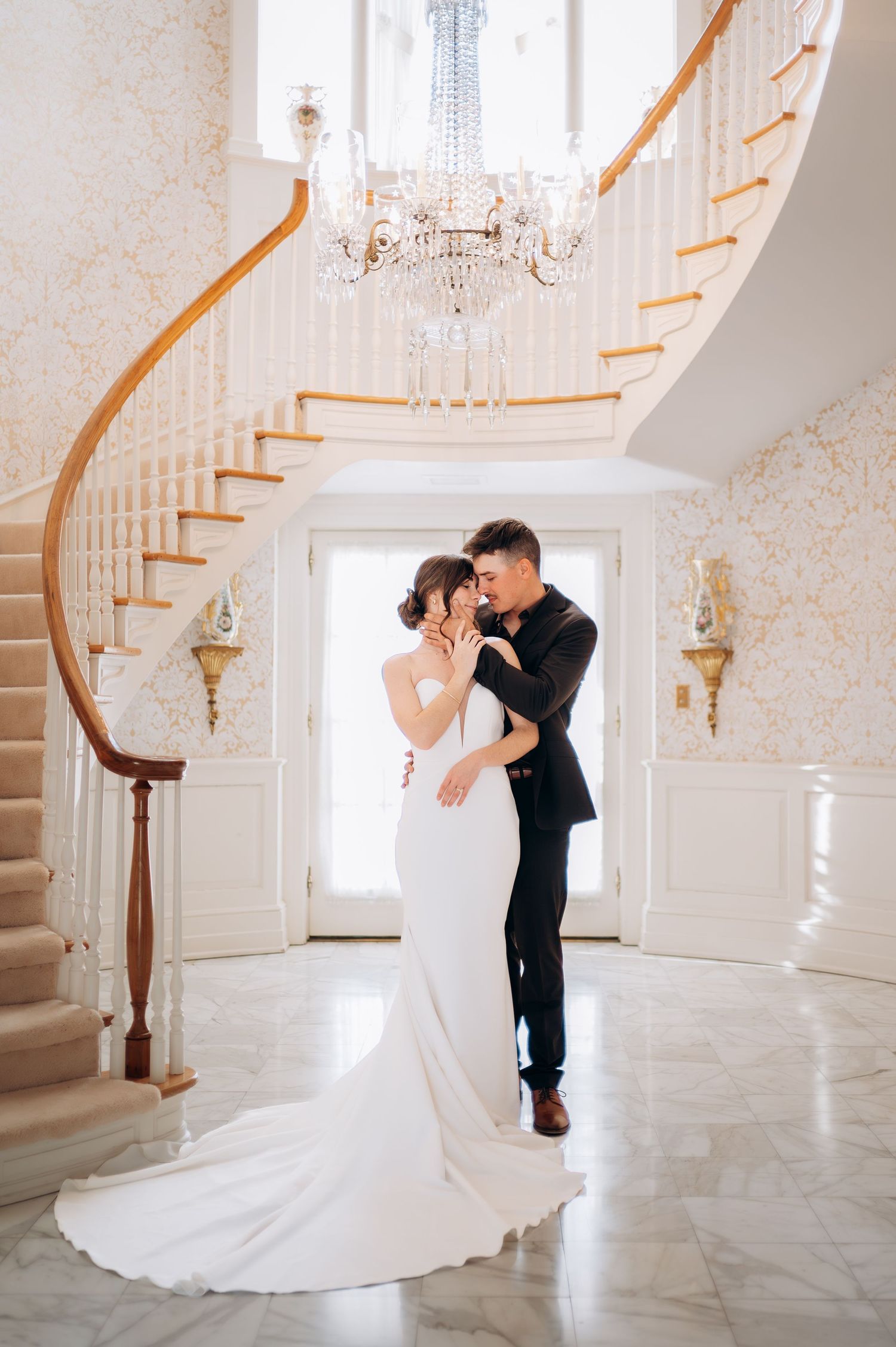 Bride and groom embrace beneath a crystal chandelier at the base of a stunning curved grand staircase.