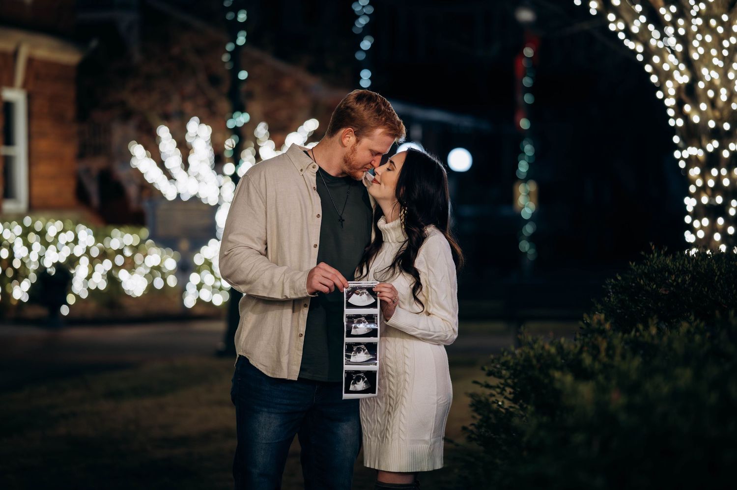 Couple sharing a tender moment holding ultrasound photos surrounded by sparkling Christmas lights.