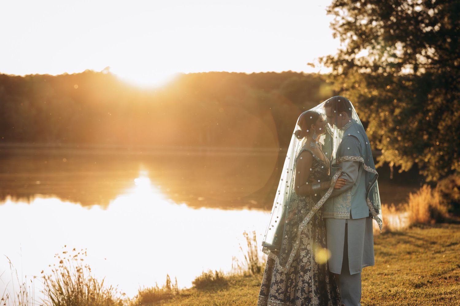 Couple sharing romantic sunset kiss by lakeside, bride in ornate veil and gown, groom in light suit, golden hour glow.