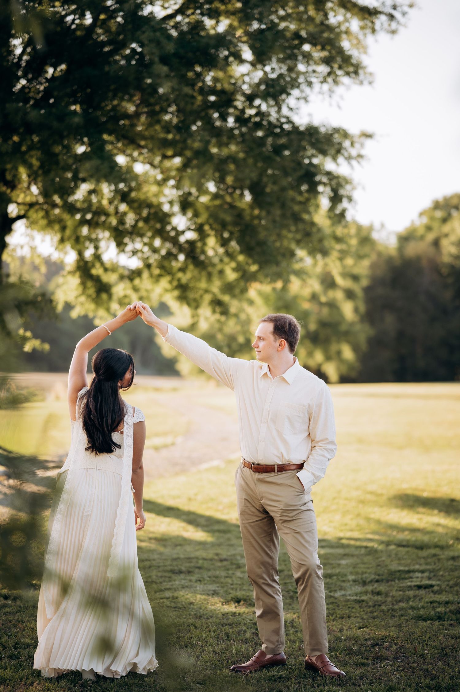 Couple dancing outdoors at golden hour, man spinning woman in white dress on lush green lawn near trees.