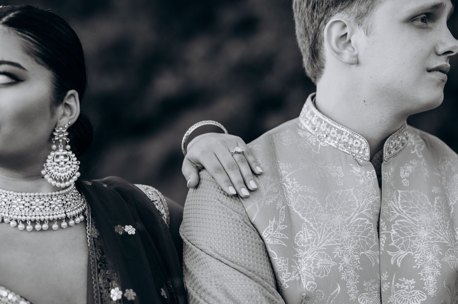 Black and white photo of a bride in Indian bridal jewelry resting her hand on a groom in an embroidered sherwani.