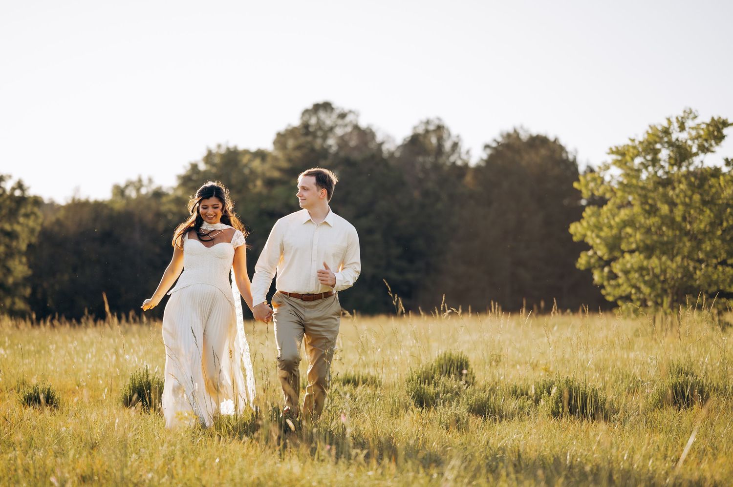 Couple holding hands walking through golden meadow in wedding attire, sun-drenched field with trees in background.
