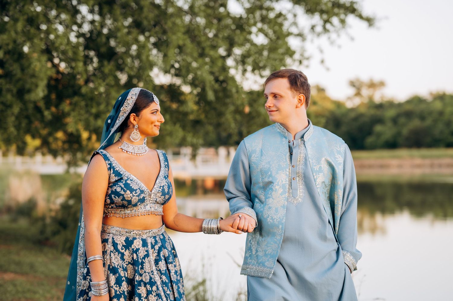 Couple in traditional Indian wedding attire holding hands outdoors near a lake, bride in blue lehenga and groom in kurta.