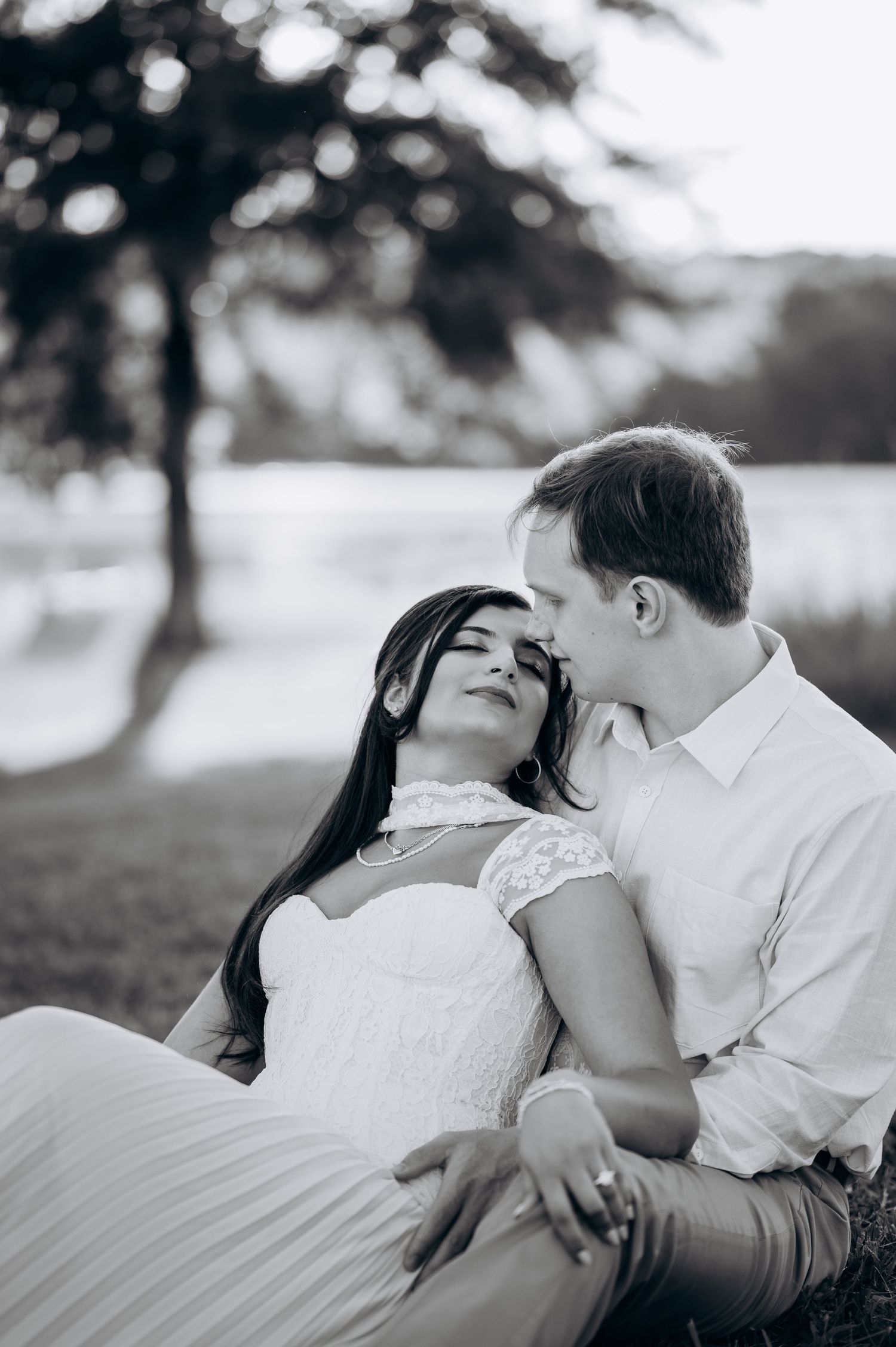 Black and white photo of a couple in wedding attire sitting together outdoors near a lake, the bride leaning back into groom.