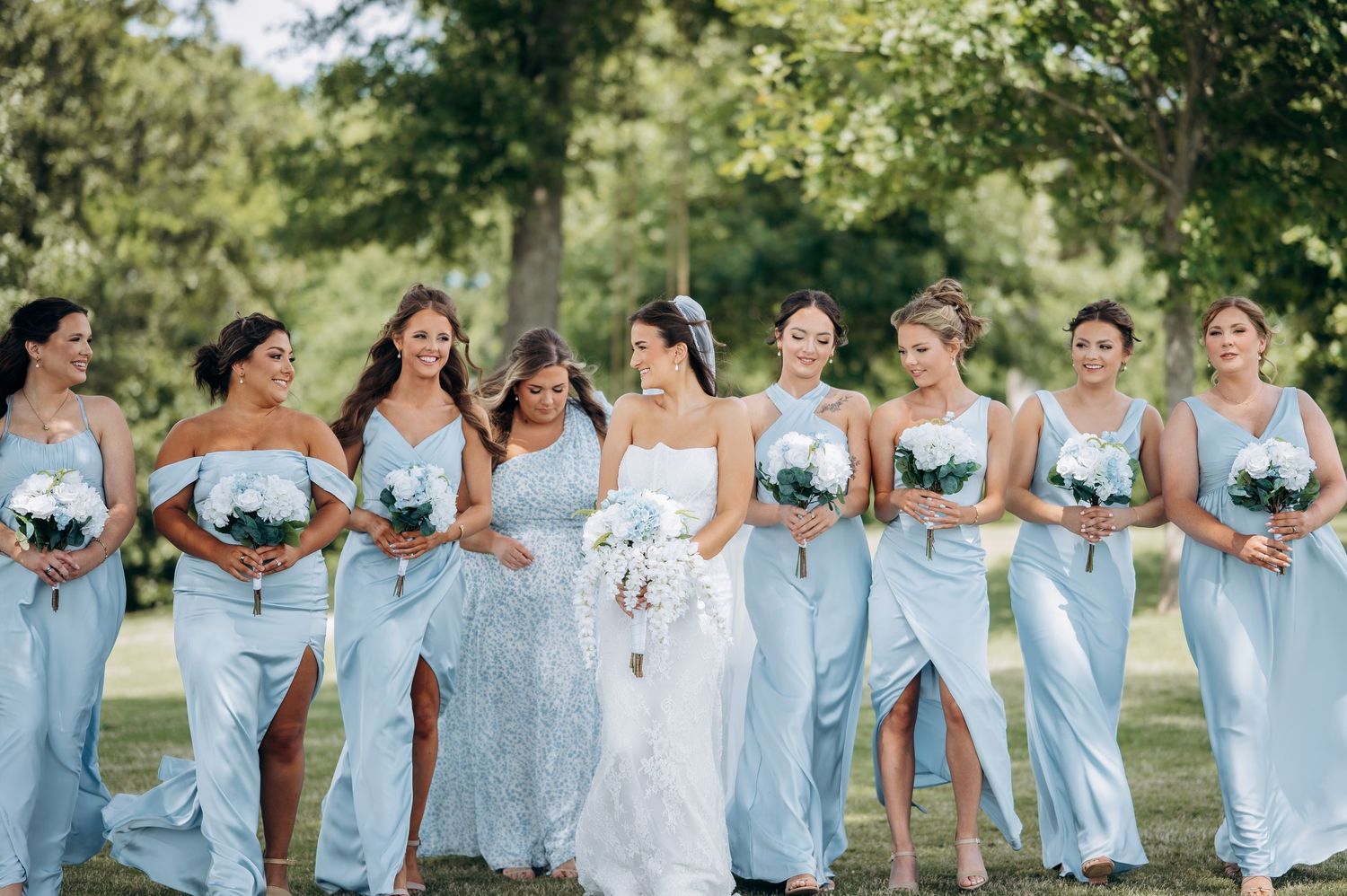 Bride in white lace gown walks with bridesmaids in light blue dresses holding white bouquets outdoors.