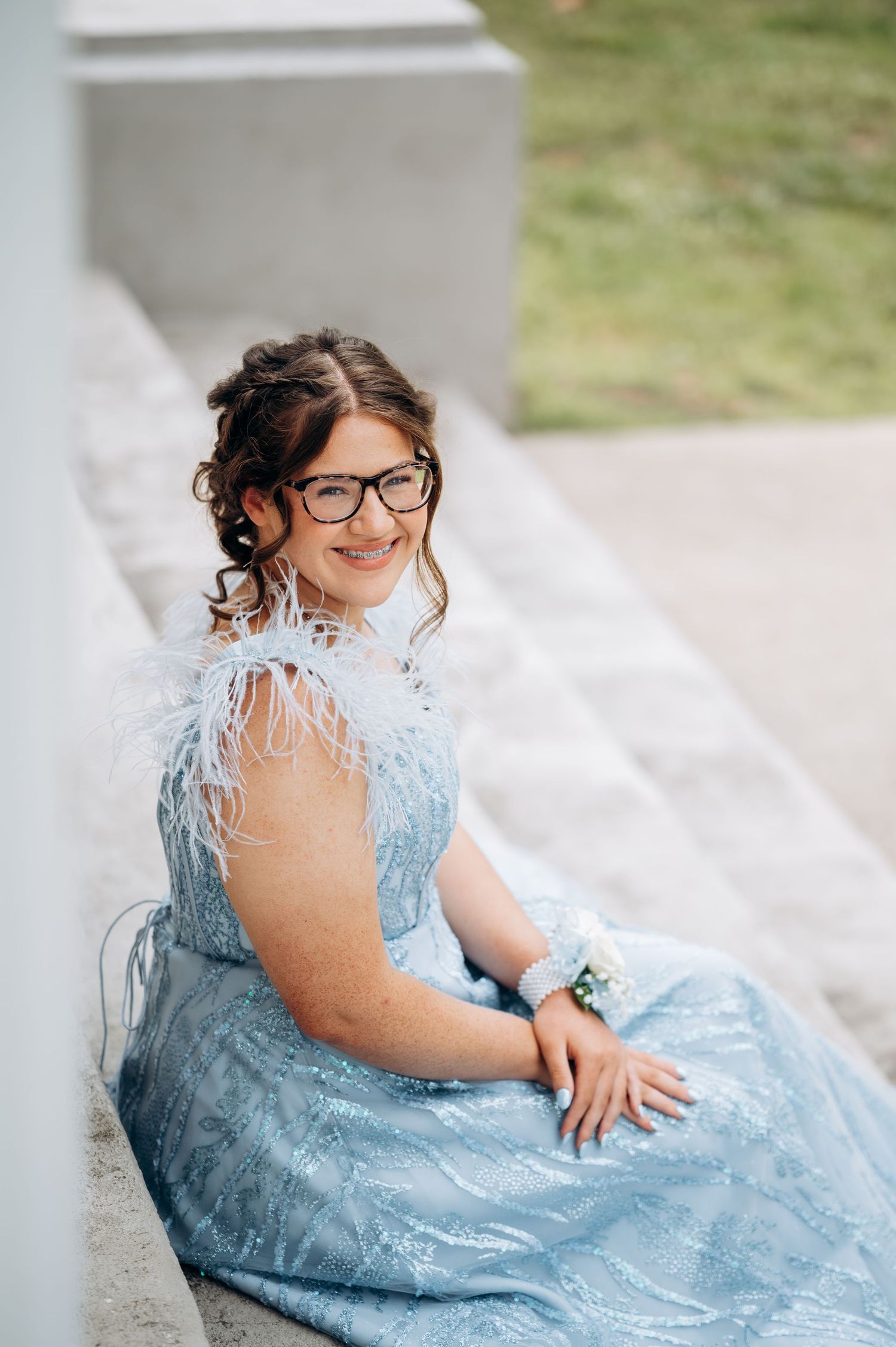 Young woman in blue sequin feather dress with wrist corsage and glasses smiling at prom.