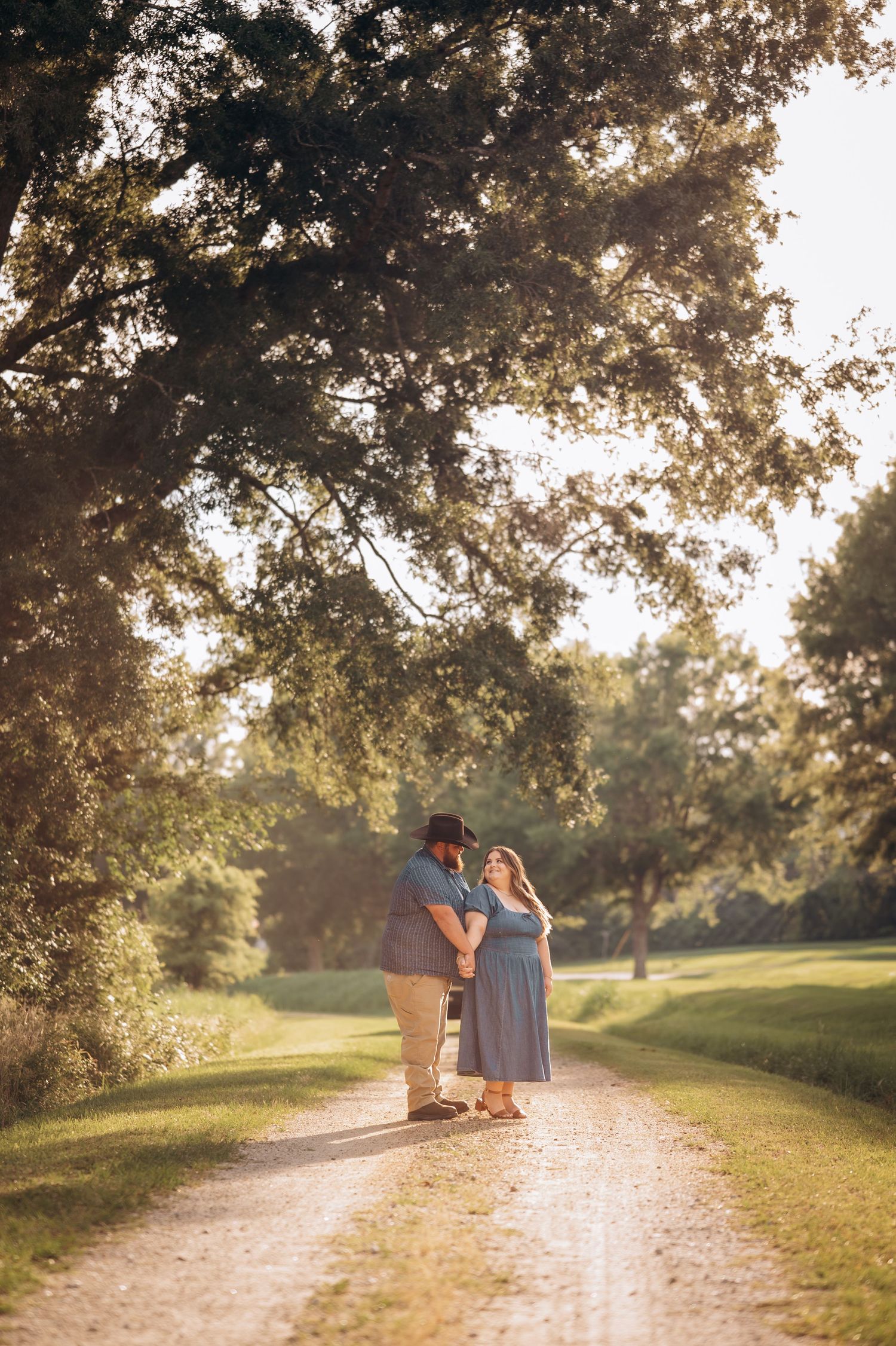 Couple holding hands on a sunlit dirt path surrounded by lush green trees during a golden hour outdoor photo session.