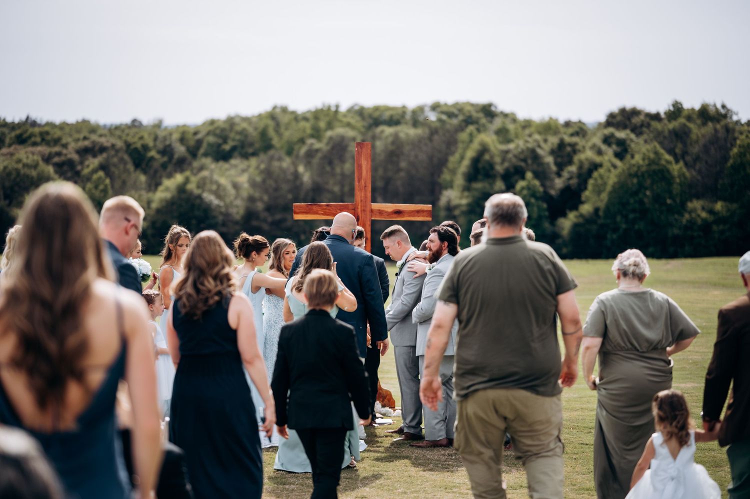 Wedding guests gathering around wooden cross at outdoor rustic field ceremony.