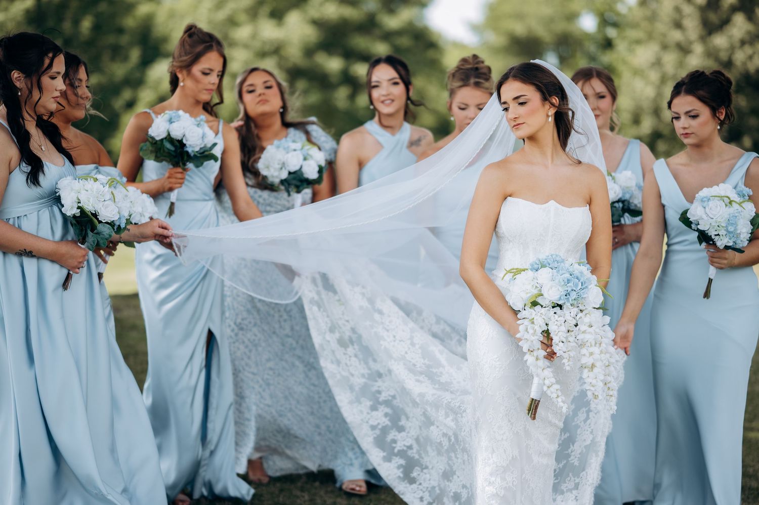 Bride in white lace gown with flowing veil held by bridesmaids in light blue dresses holding white bouquets outdoors.