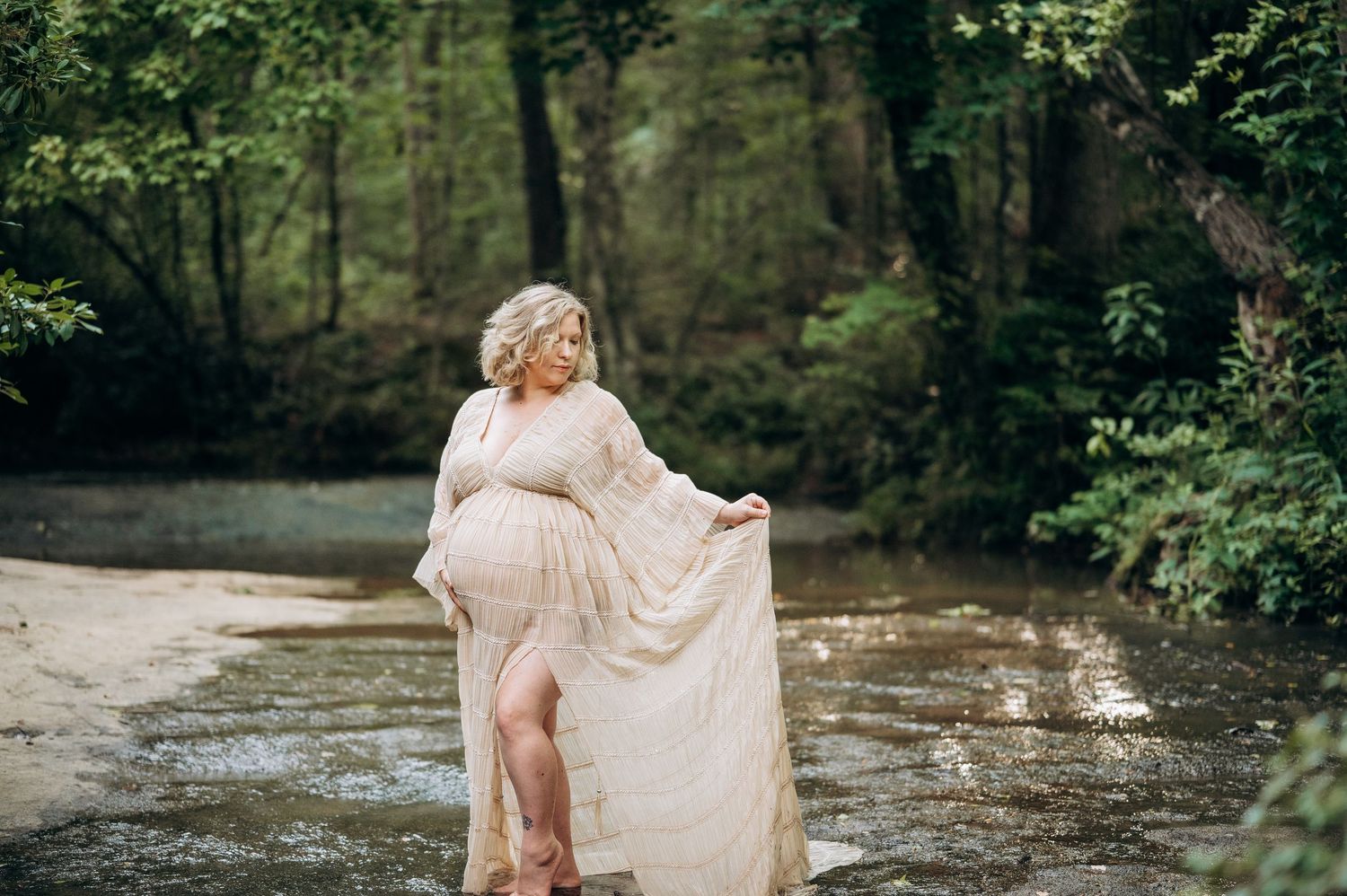 Pregnant woman in flowing lace gown standing in a forest creek, showcasing maternity elegance outdoors.