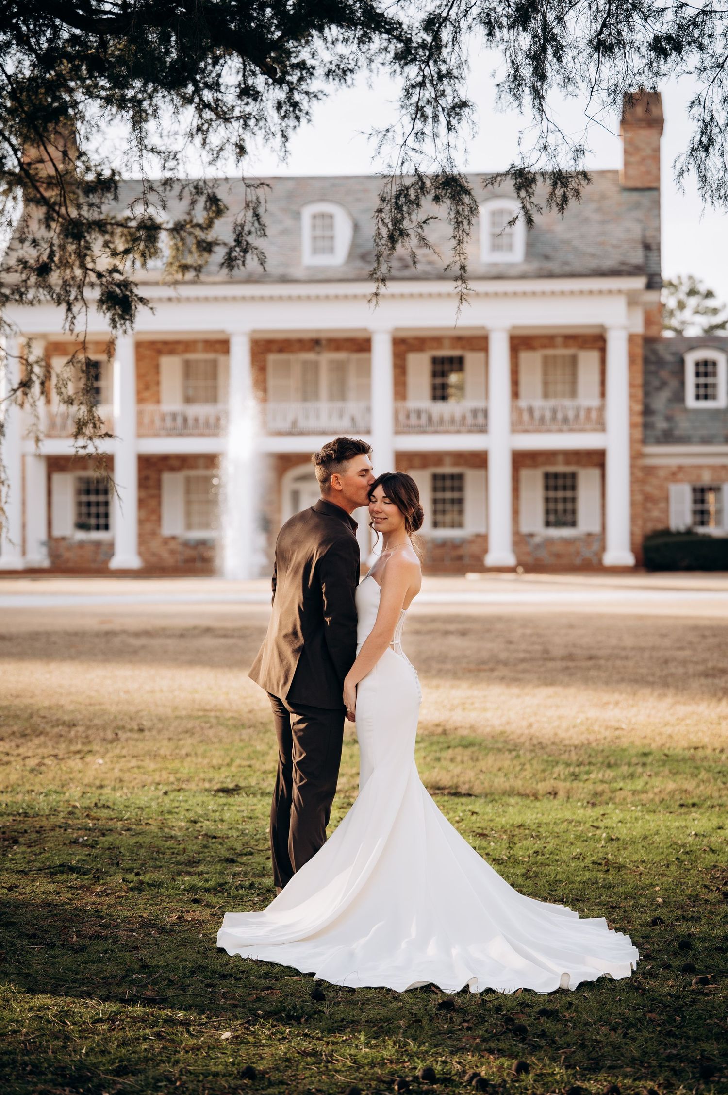 A wedding couple poses together on a lawn in front of a grand historic colonial-style building at sunset.