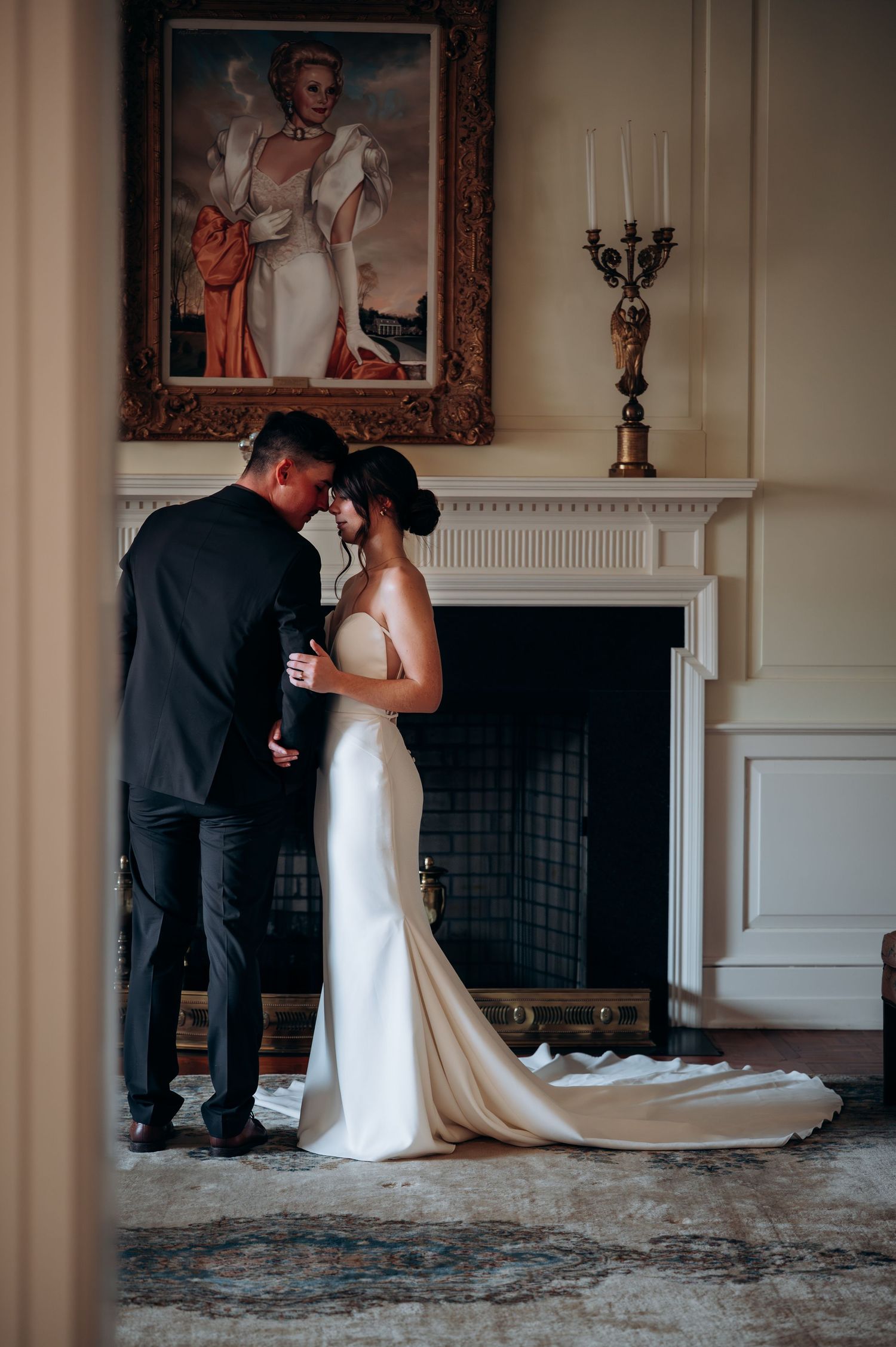 Bride and groom standing together before fireplace beneath classic portrait painting with candelabra accent.