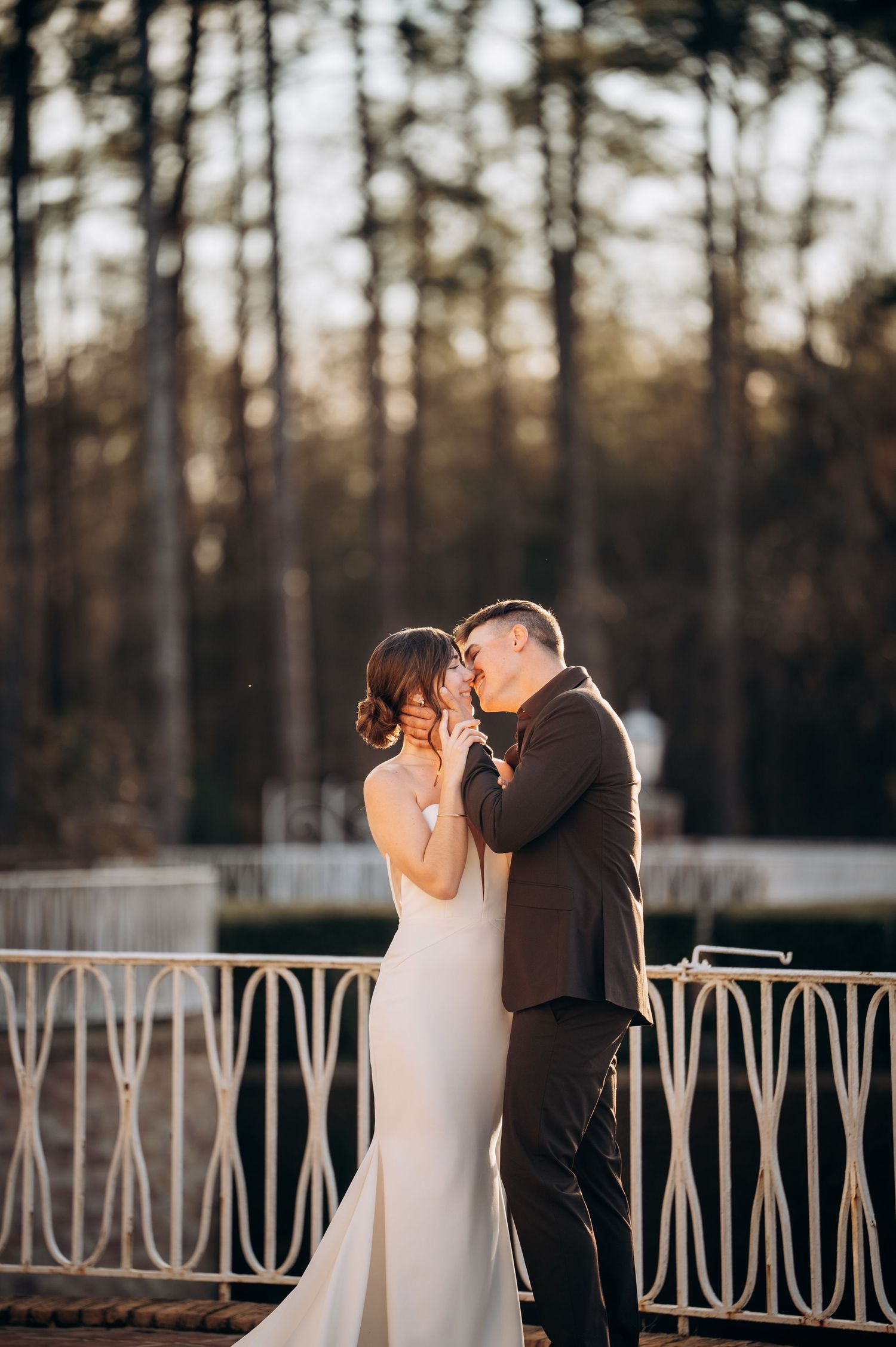 Bride and groom share a kiss on a decorative white railing bridge with a sunlit forest backdrop.