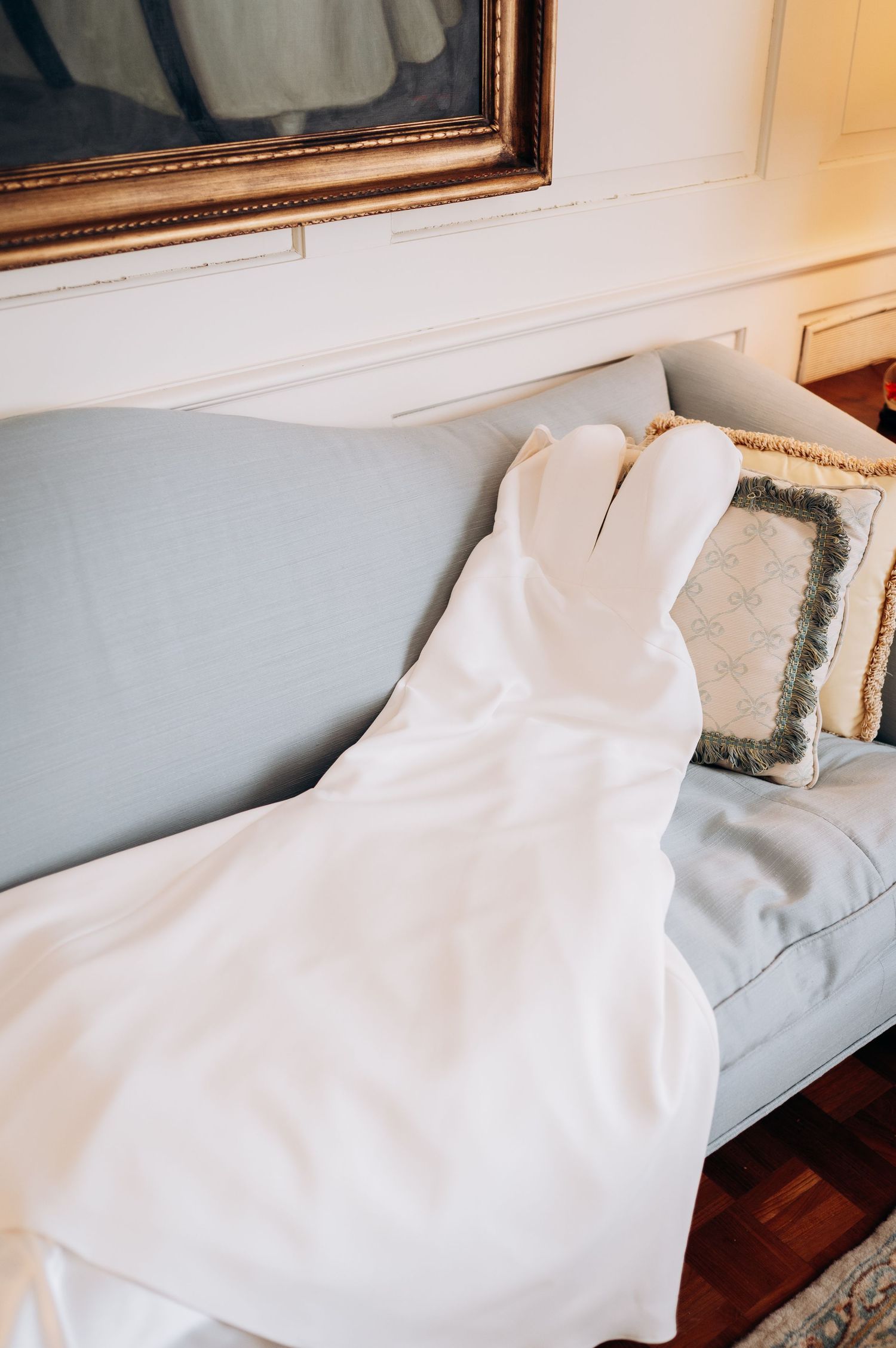 White wedding dress draped over a light blue sofa near a window with a decorative pillow.