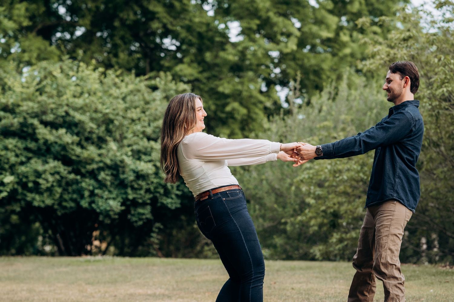 Happy couple holding hands and spinning together in a sunny park with green trees in the background.