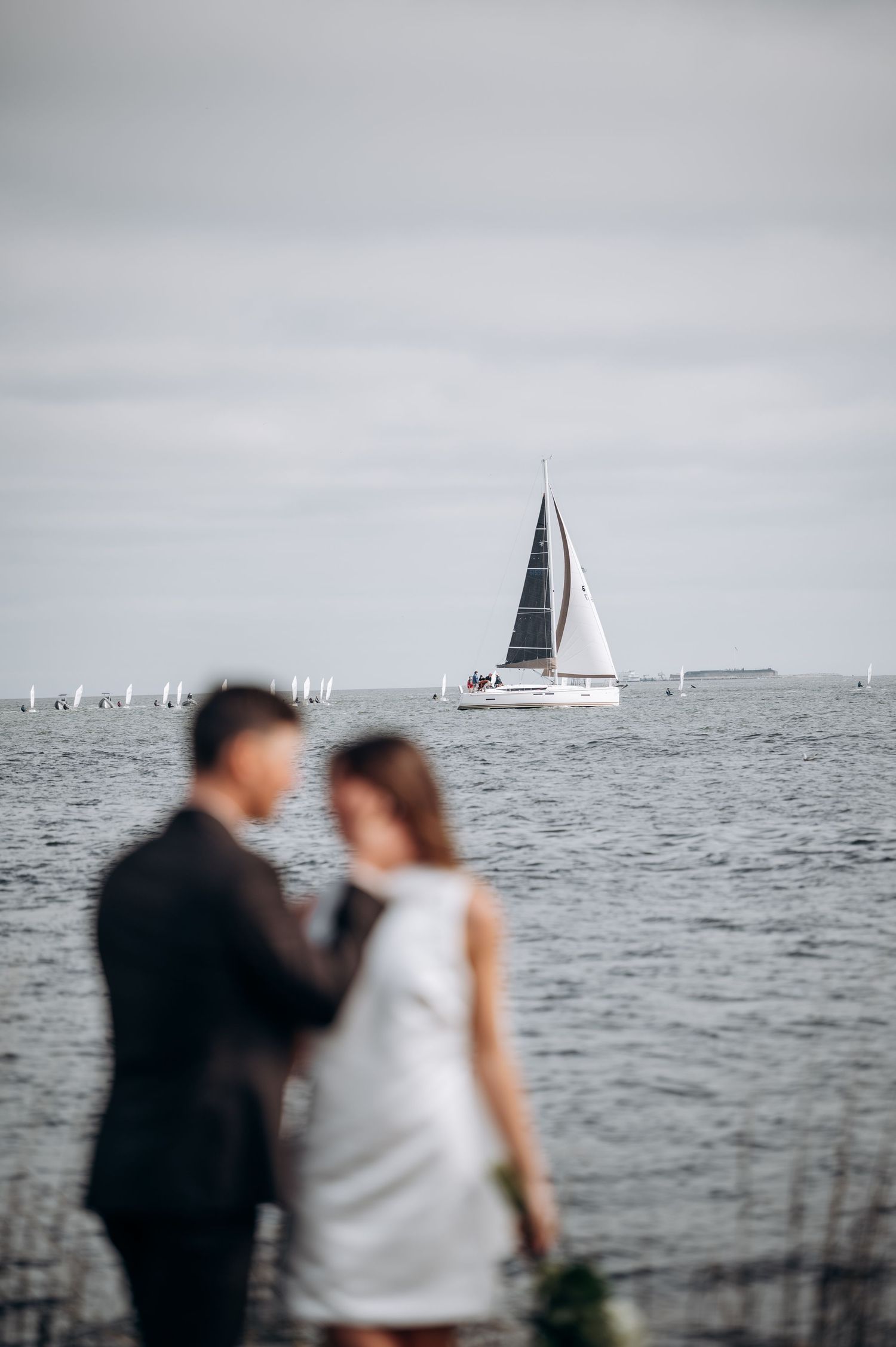 Blurred newlywed couple standing romantically before a serene harbor with a sailboat in the background.