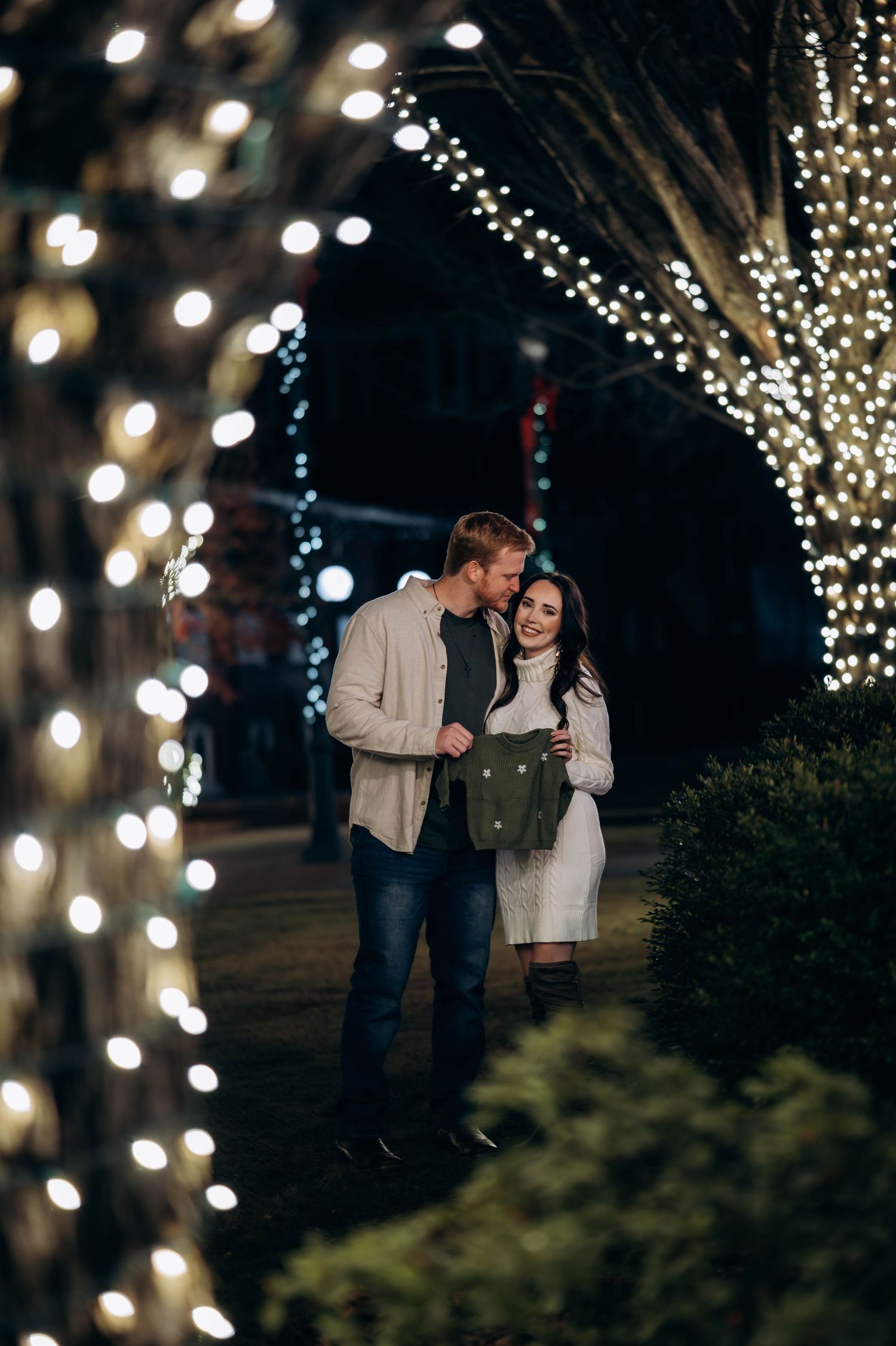 Couple embracing among twinkling Christmas lights on trees at night, woman smiling in white dress.