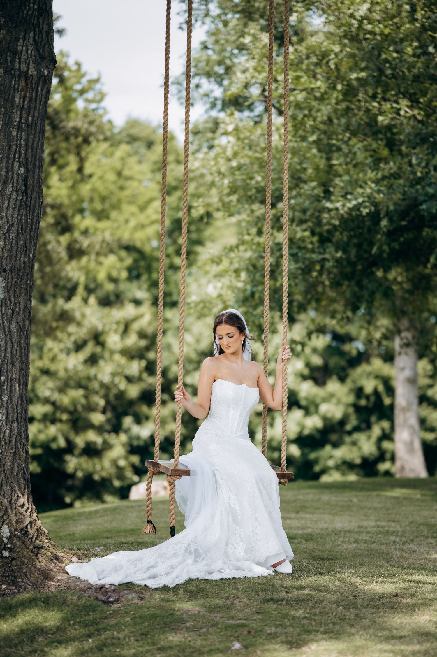 Elegant bride in white strapless wedding gown sitting on a rope swing surrounded by lush green trees.